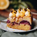 A close up reveals the layers of a piece of plum and Lamond cake. Black doris plums are seen in the centre and lemon curd and cream is piped on top of the slice with yellow flower petals on top.