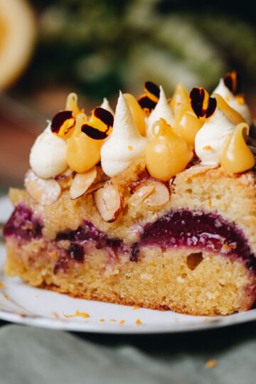 A close up reveals the layers of a piece of plum and Lamond cake. Black doris plums are seen in the centre and lemon curd and cream is piped on top of the slice with yellow flower petals on top.