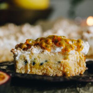 A close up shot shows a piece of passionfruit lemon meringue slice on a black plate. Fresh passionfruit is added on top of the meringue topping.