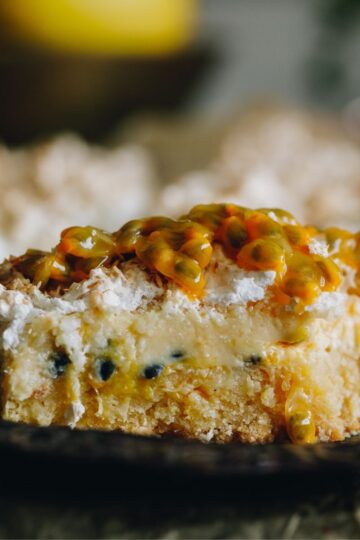A close up shot shows a piece of passionfruit lemon meringue slice on a black plate. Fresh passionfruit is added on top of the meringue topping.
