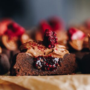 A close up reveals a chocolate and raspberry cookie that has been cut in half to reveal the raspberry jam inside of the chocolate biscuit with chocolate buttercream and a raspberry on top.
