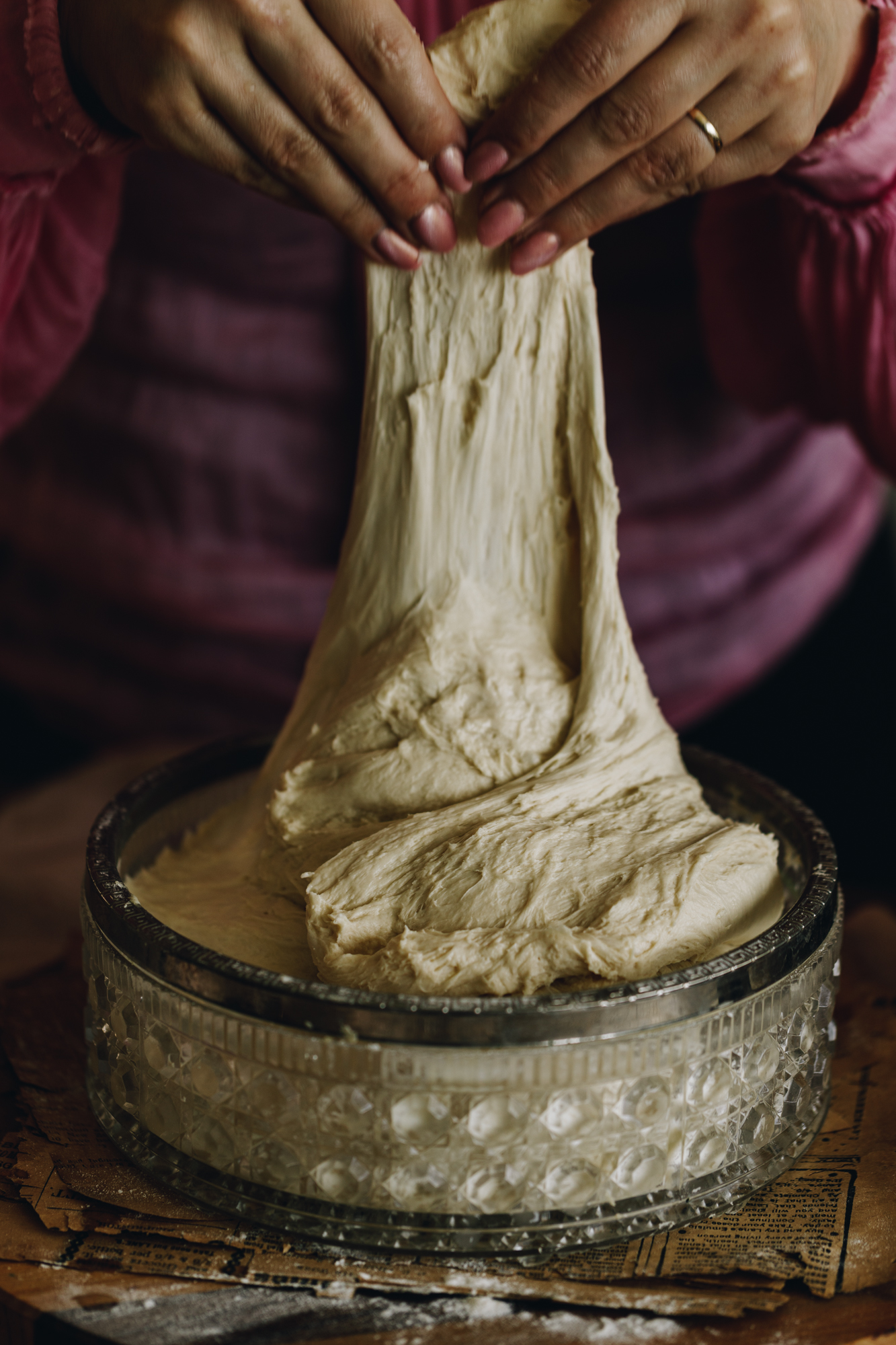 No-knead bread bun dough is being stretched and folded. The dough is being lifted high out of the vintage glass bowl. It is on a wooden board.