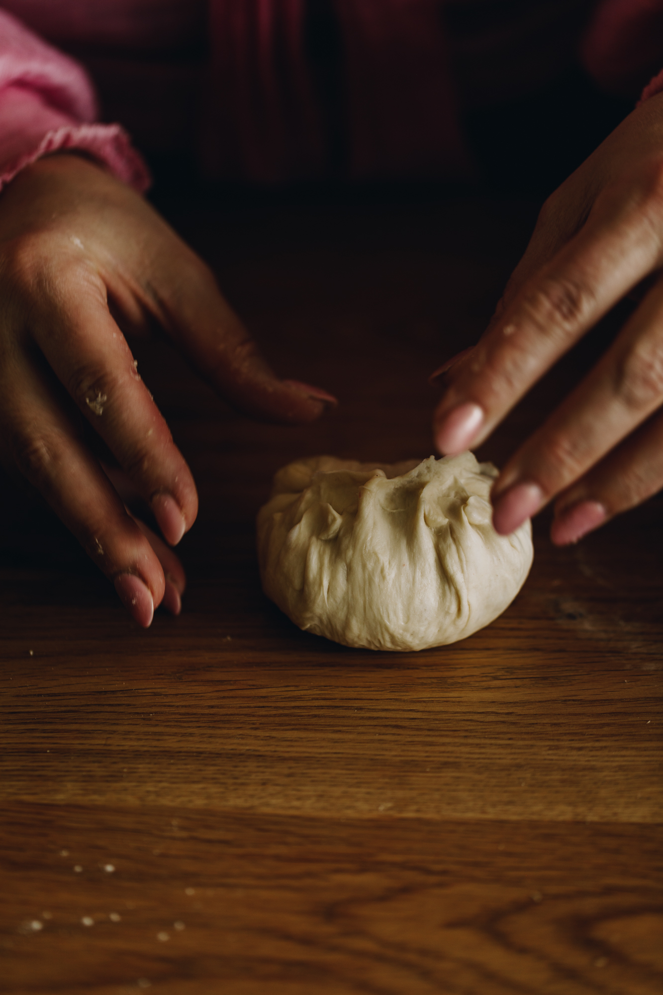 On a wooden table, hands are shaping a piece of dough in to bun, the seams have been pinched. 