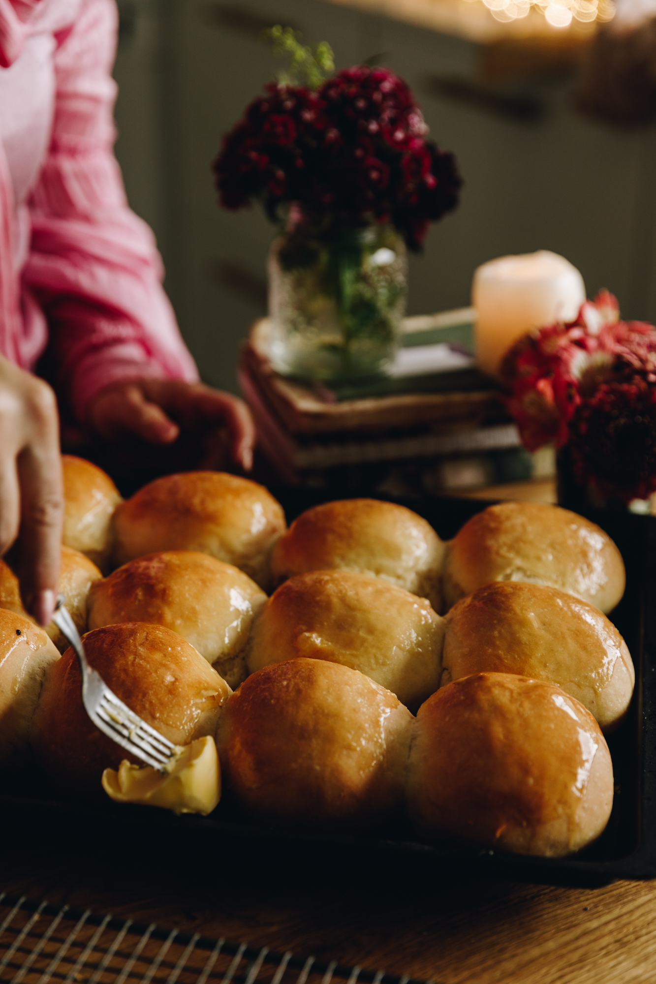Freshly baked no-bake-bread-buns are on a black tray are are being glazed with butter, giving them a shiny appearance. They are on a wooden table. 