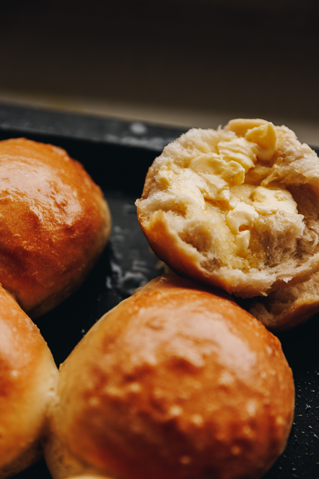 A close of freshly baked no-knead bread buns on a black tray. One of them has been cut open to reveal the fluffy centre, it has melty butter on it. 