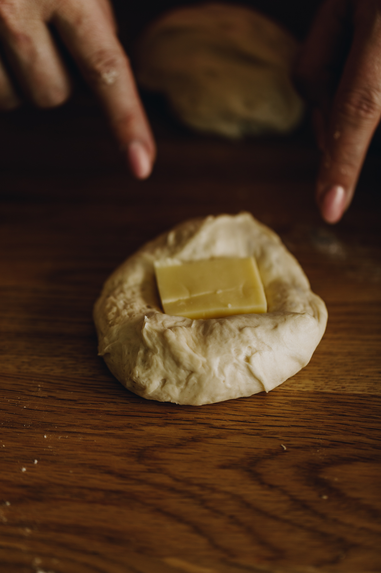 On a wooden table, hands are pressing a square of mozzarella cheese in to unbaked cheesy no-knead bread dough. 