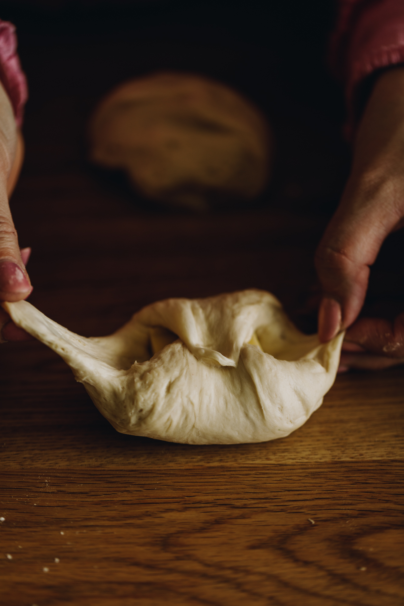 On a wooden table, hands are wrapping the square of mozzarella cheese in to unbaked cheesy no-knead bread dough.
