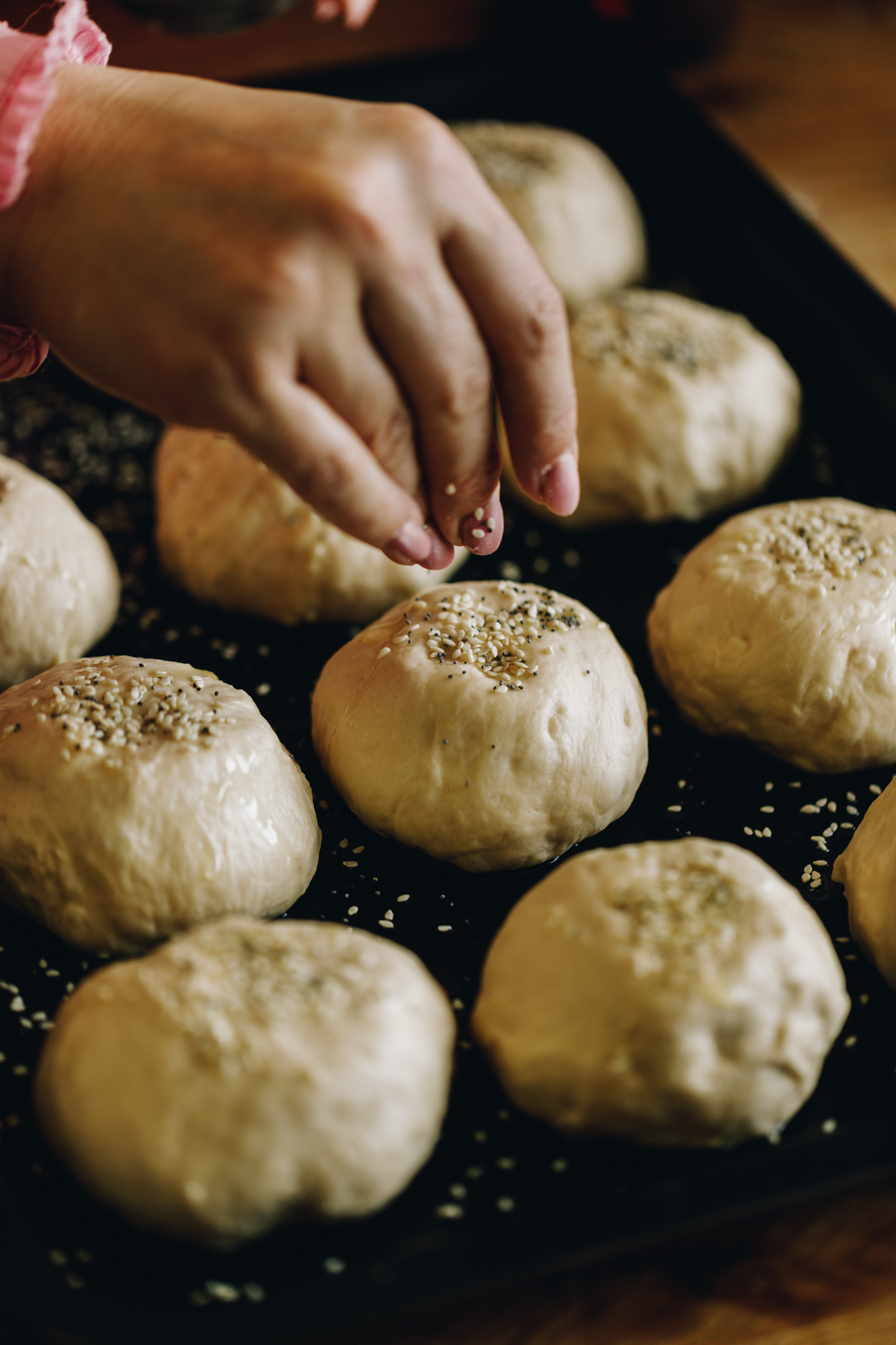 Unbaked cheese no-knead bread buns are on a black tray. They are being sprinkled with bagel seasoning on a wooden table. 