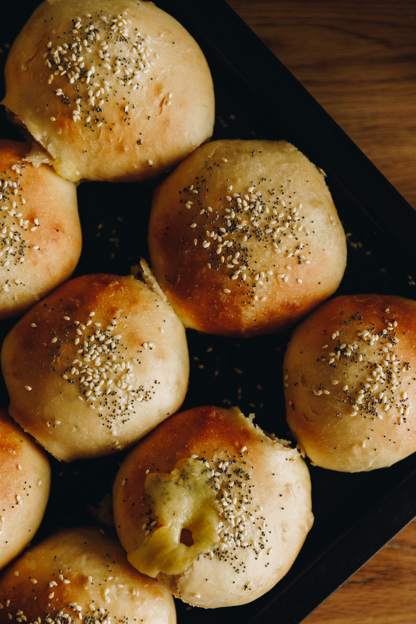A flat lay shot shows freshly baked cheesy no-bake bread buns are on a black tray, on a wooden table. 