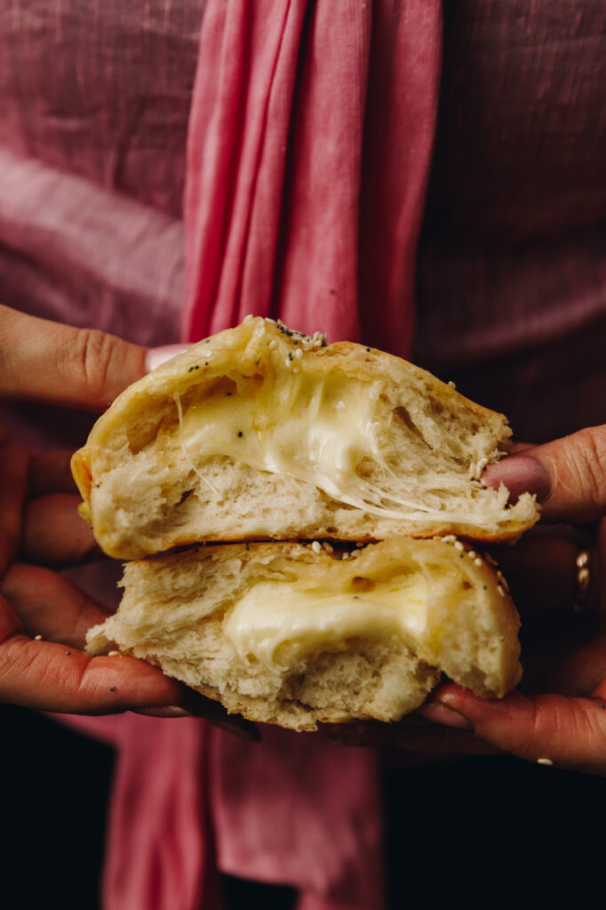 A close up shot of cheesy no-knead bread buns shows a bun cut open and the gooey cheese is oozing out of the centre. 