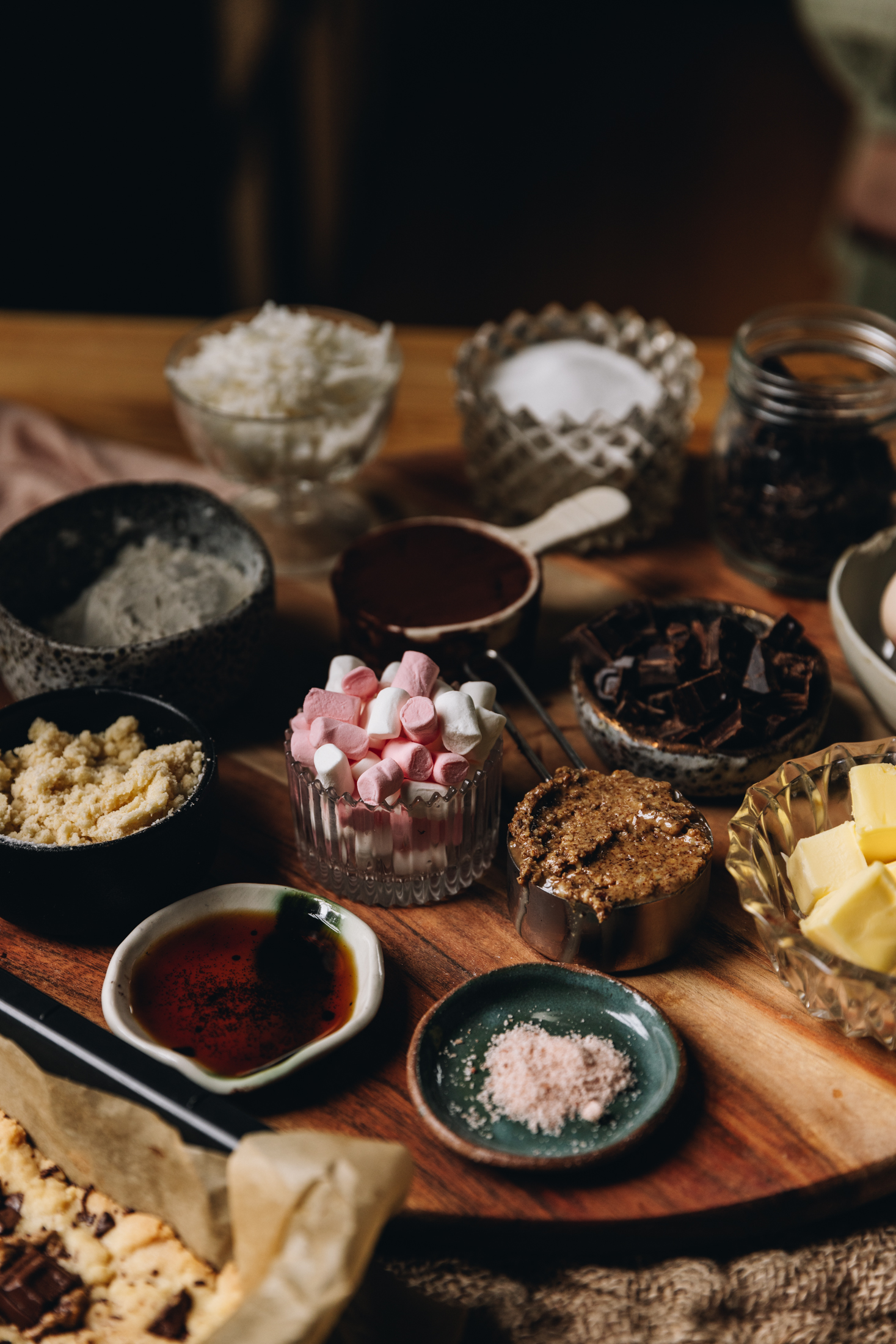 A round wooden board sits on a rustic cloth, on a wooden table. On the board is an array of small vintage plates and vases. Ingredients are added to each one, in view are salt, vanilla, mini marshmallows, almond butter, butter, chocolate, sugar and coconut. 