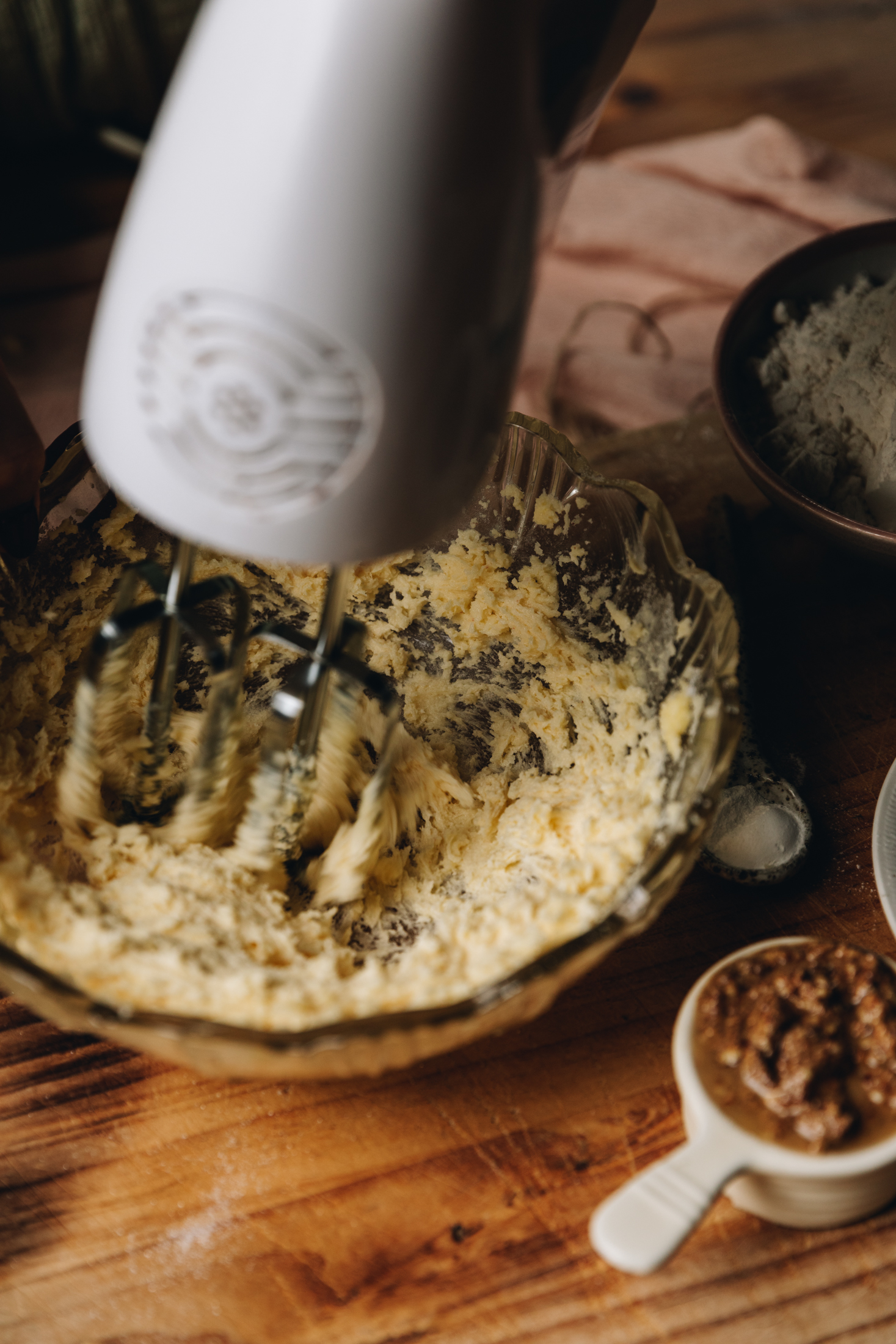 On a wooden table sits a glass vintage bowl that is has butter and sugar being whipped in ti with a white hand mixer. Next to it is a small ceramic cup that has almond butter in it. Other ingredients are in the background. 