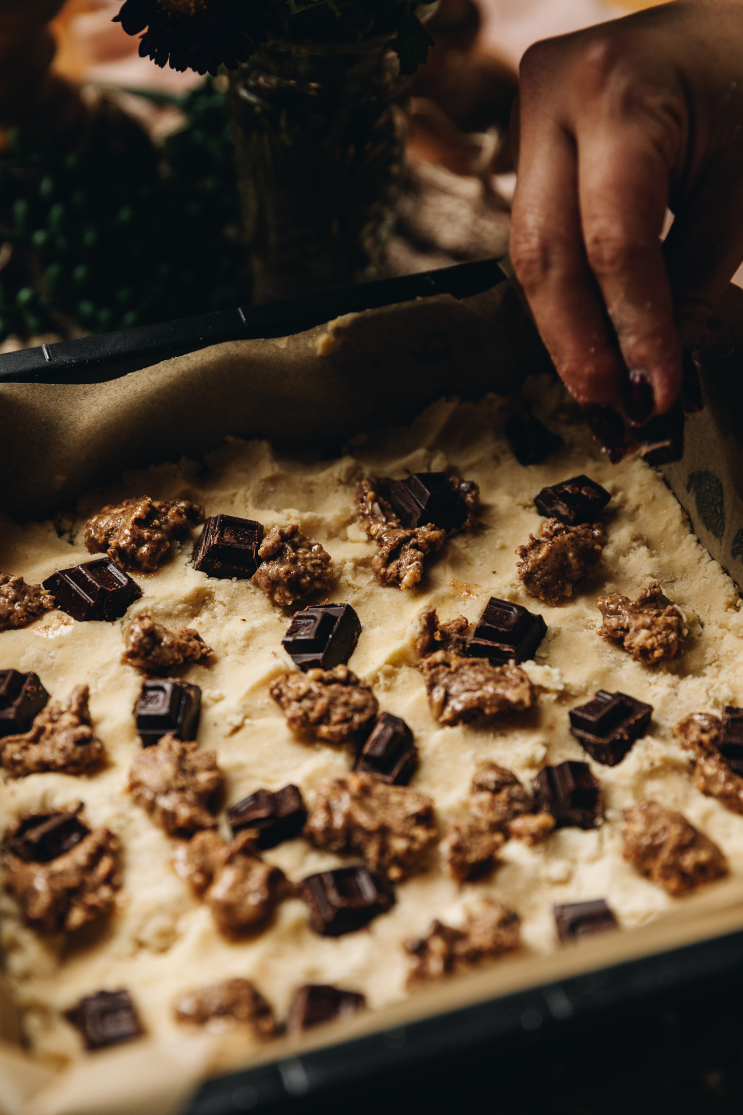 A vintage baking tin is lined with brown baking paper. In it is the biscuit base for Brookie slice. It has chopped dark chocolate and dots of almond butter all over it. Naomi is placing another piece of chocolate on to the base. 