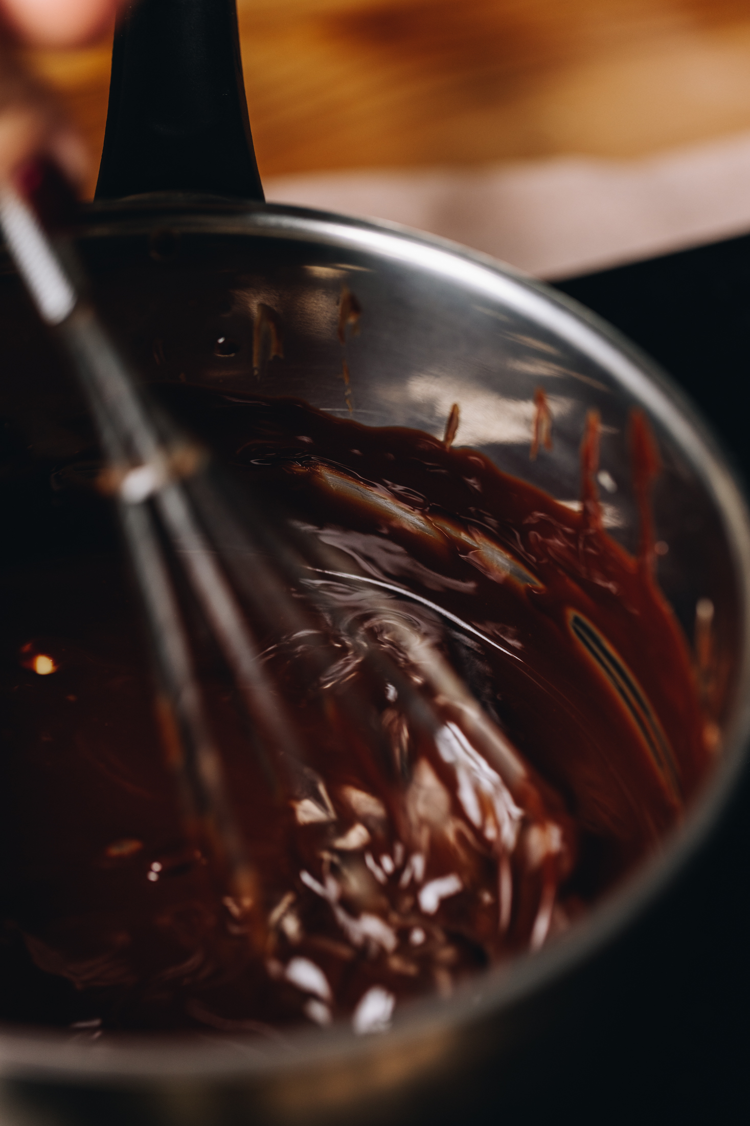 A silver pot with a black handle sits on a black portable stove. In the pot is melted dark chocolate with a wire whisk in it too.  A wooden table is seen in the background.