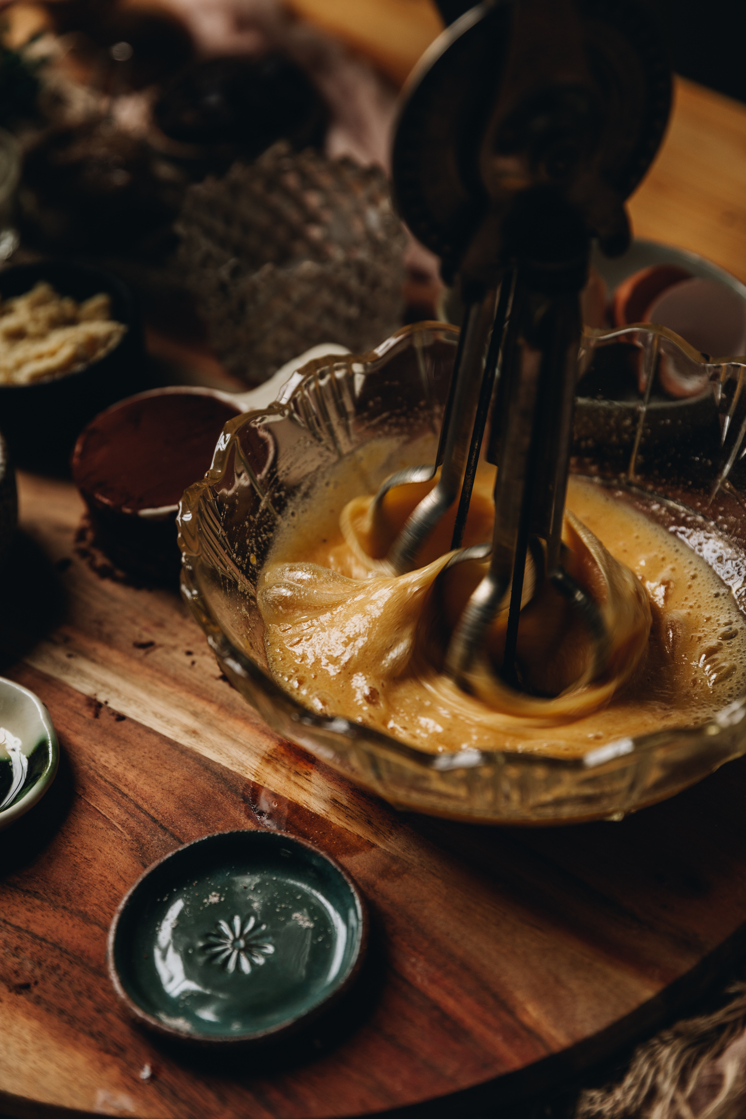 On a round wooden board sits a vintage glass bowl. In it is eggs that are being whipped by a vintage egg beater. Next to the bowl is a tiny green plate and a white ceramic cup with cocoa in it. Other vintage jars are in the background. 