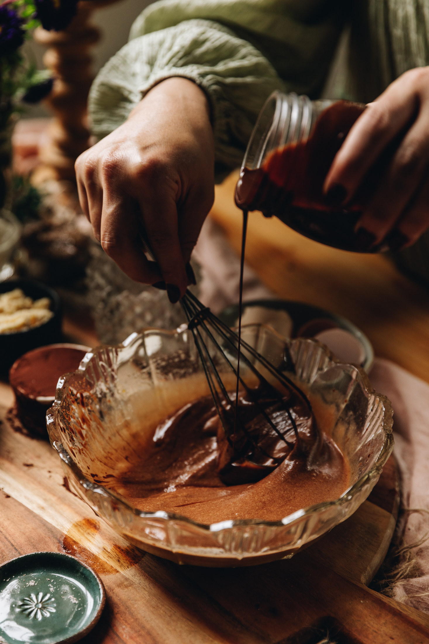 On a round wooden board sits a vintage glass bowl. In it is eggs that are being whipped by a wire whisk as melted chocolate is being poured in to it. Next to the bowl is a tiny green plate and a white ceramic cup with cocoa in it.
