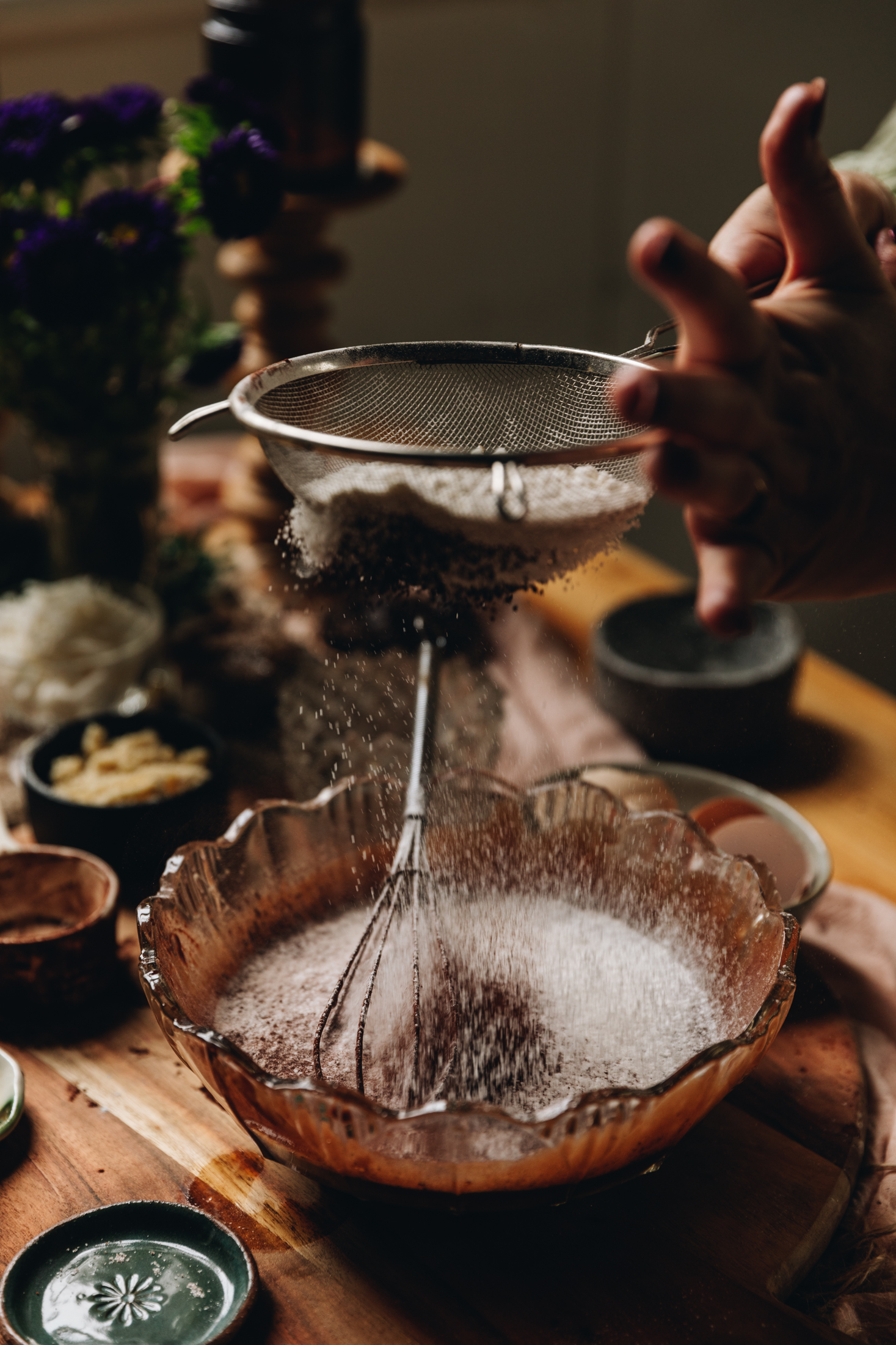 On a round wooden board sits a vintage glass bowl. In it is a chocolate mixture. Above the bowl is a wire sifter that is sifting flour and cocoa in to the bowl. Next to the bowl is a tiny green plate and a white ceramic cup with cocoa in it. There is several vintage bowls in the background and a wooden candle holder with purple flowers next to it. 