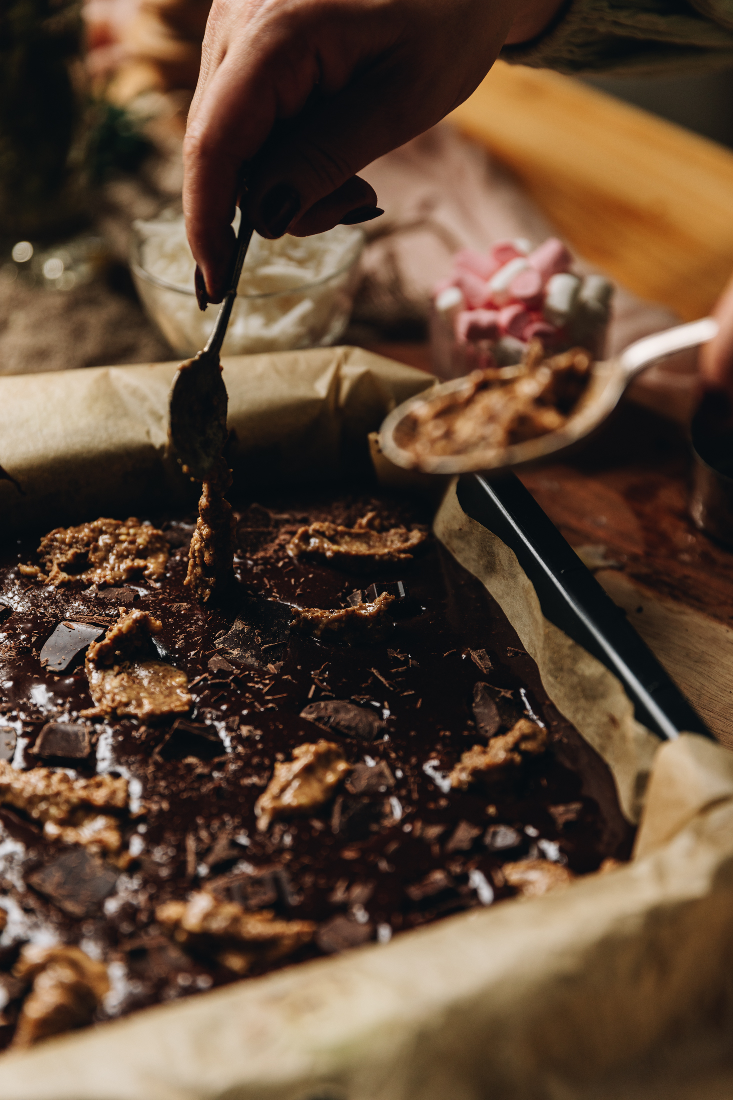 A vintage baking tin is lined with brown baking paper. In it is the unbaked brownie part of the brookie slice. It has chunks of chocolate and almond butter all over the top. Naomi is adding more nut butter with a teaspoon and it is dripping off. A wooden table is in the background.