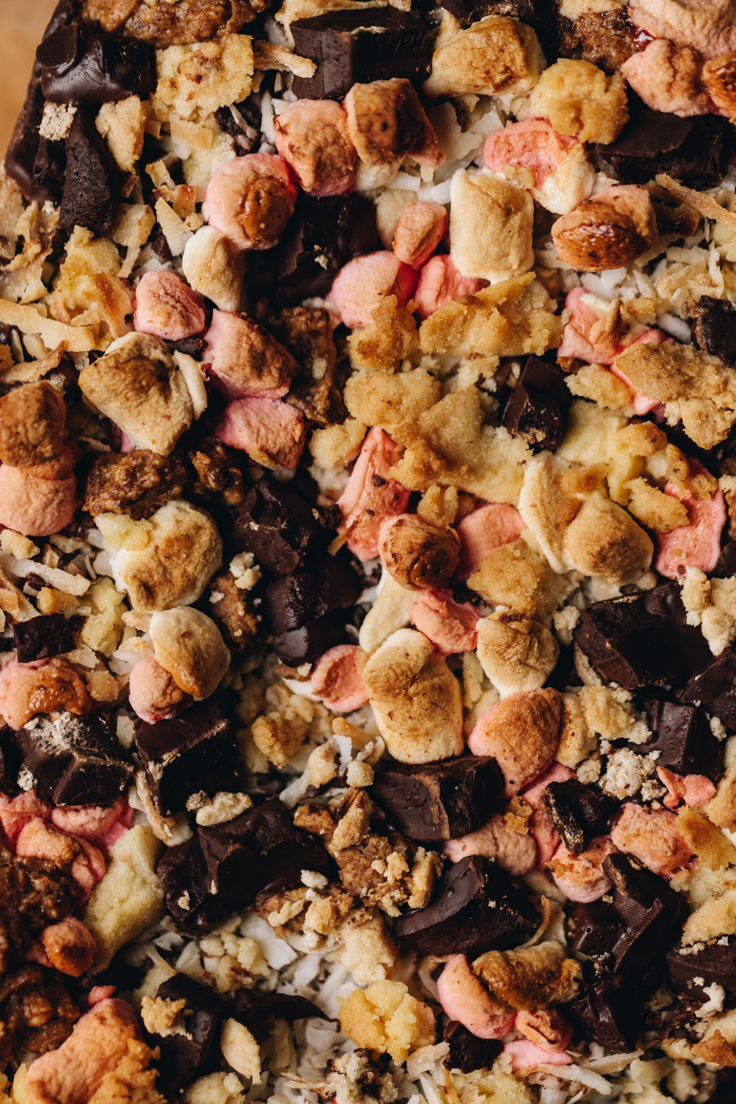 A close up shot reveals the brookie slice topping that has been baked. There is chunks of dark chocolate, shredded coconut, cookie crumbs and toasted pink and white marshmallows.