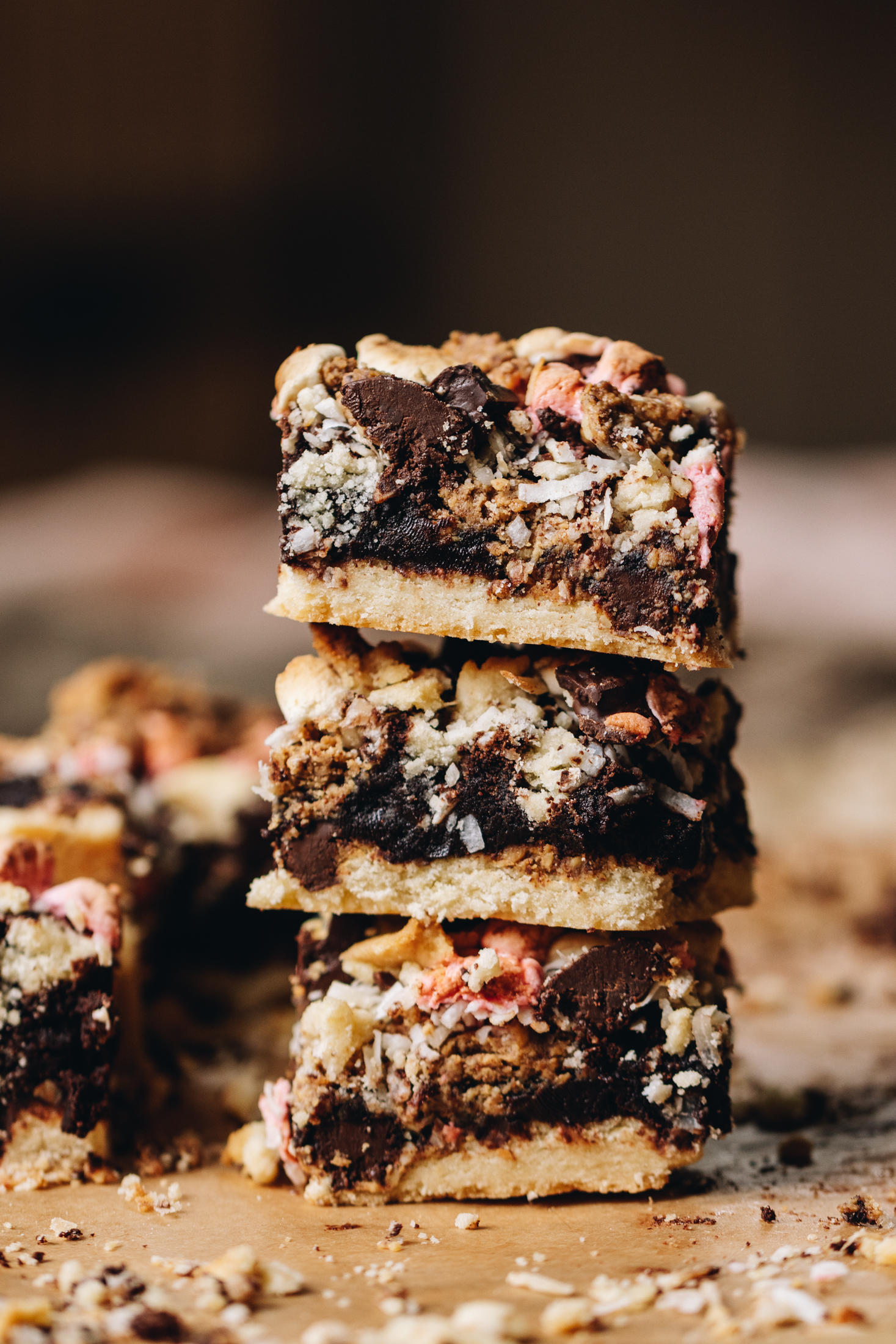 Three pieces of Brookie slice have been stacked on top of each other. The shot reveals the cookie base, the brownie centre with peanut butter and chocolate and the marshmallow topping. There are coconut and cookie crumbs all over the board that the brookie slice is on.