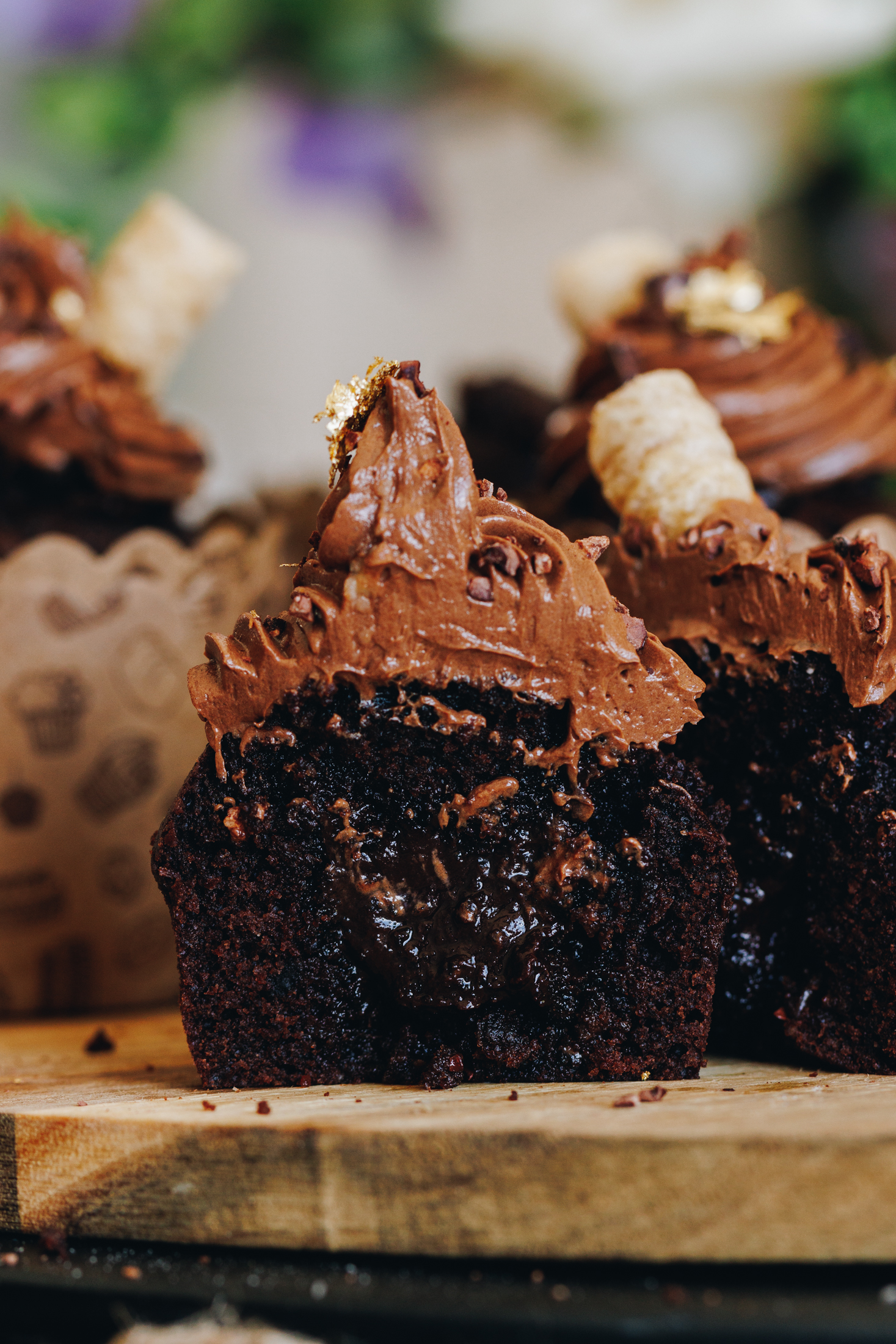 Chocolate ganache  muffins sit on a wooden board. One of the muffins is in focus and it has been cut in half to reveal the gooey chocolate centre and the chocolate frosting piled on top. 