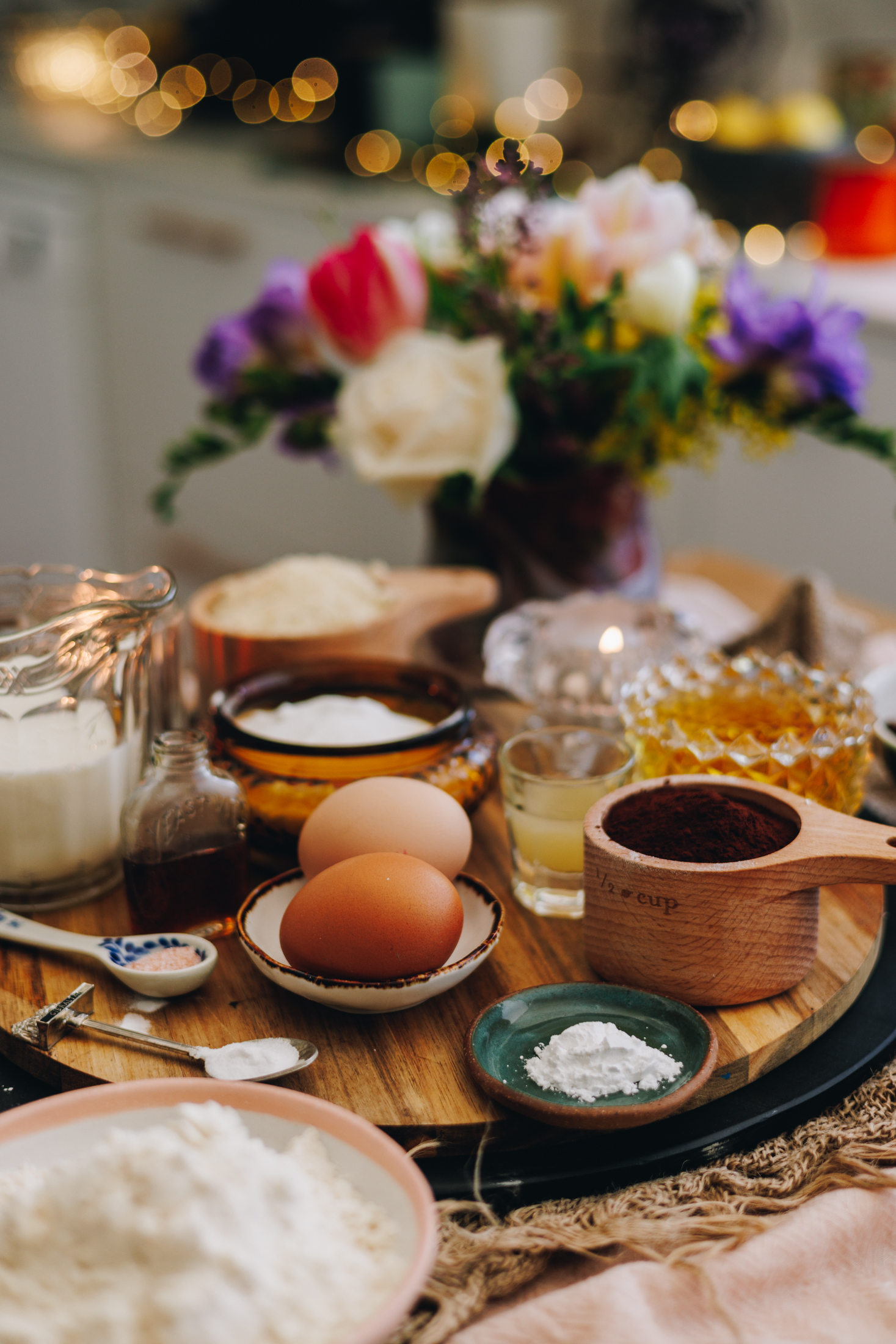 A round wooden board is on a table, on top of natural fabric and a pink tablecloth. On the board is muffin ingredients that are in vintage spoons, plates, cups and glasses. There is eggs, cocoa, baking powder, salt, milk, ground almonds, flour and oil in view. A candle burns behind the ingredients and a glass jar with flowers is behind the candle. Golden fairy lights burn in the background.