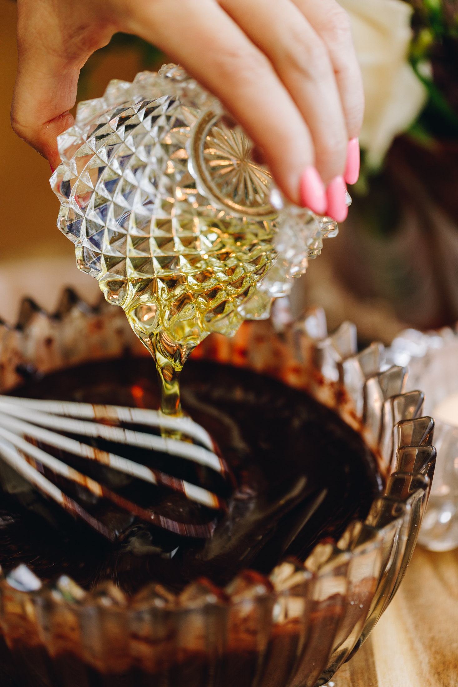 A vintage glass bowl has a thick cocoa mixture in it. Naomi is stirring it with a white whisk as she pours in oil from a vintage glass. Naomi is stirring it with a white whisk.