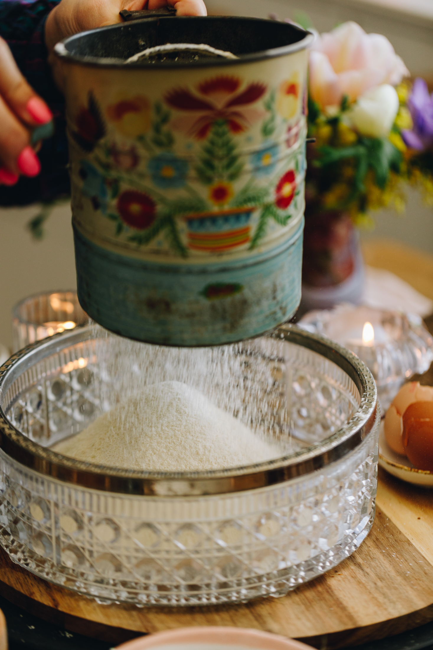 A glass vintage bowl with a silver trim sits on a wooden round board. Above the bowl is a vintage blue sifter with a wooden handle that Naomi is using to sift the dry ingredients in to the bowl. There is a candle and a bunch of flowers in the background.