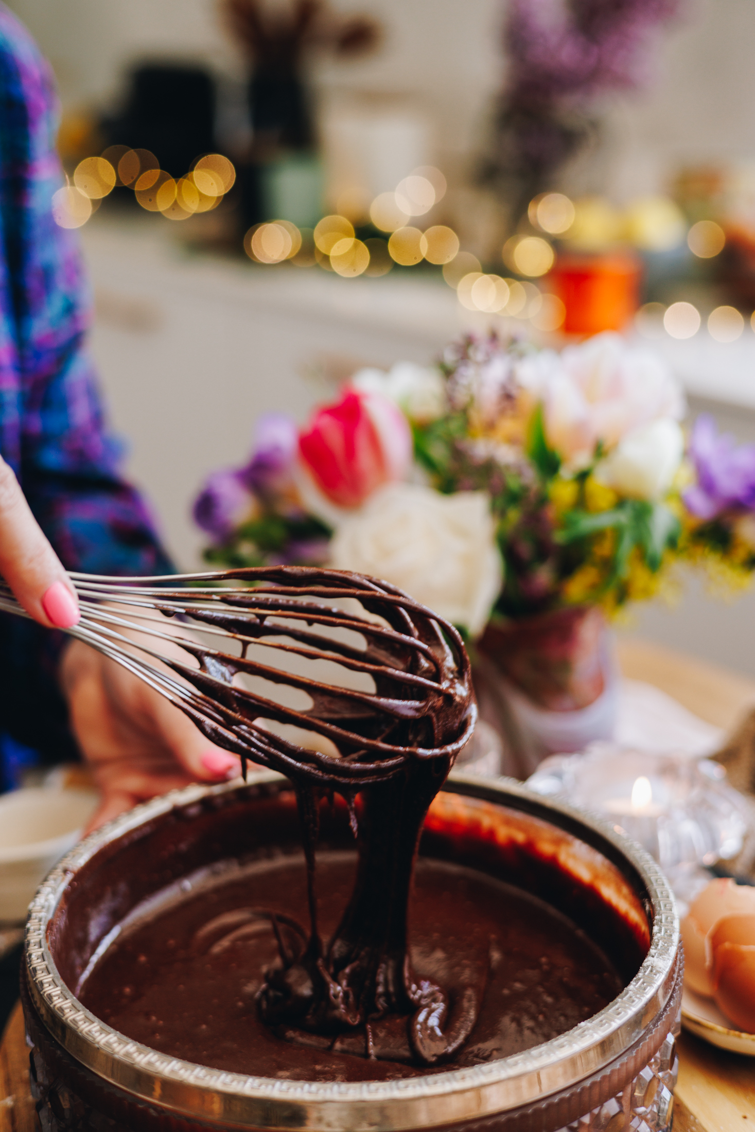A glass vintage bowl with a silver trim sits on a wooden round board. A thick chocolate mixture is in the bowl and a whisk is lifted out of the mixture with it dripping off in to the bowl. Flowers are in the background with blurred fairy lights in the background.