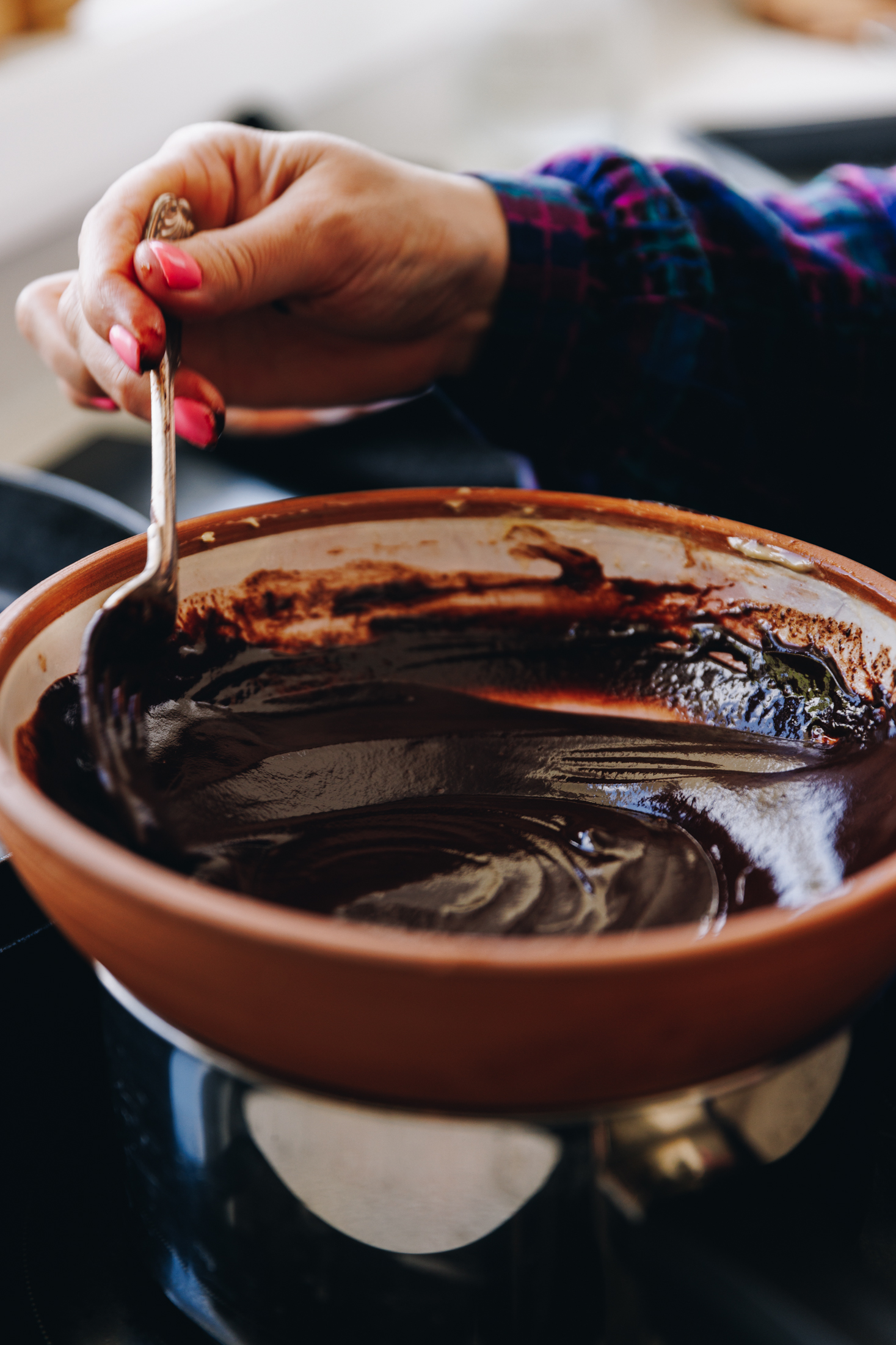 A ceramic bowl sits on a silver pot on a black stove top. In the bowl is a melted chocolate ganache mixture that is being stirred by Naomi with a fork.