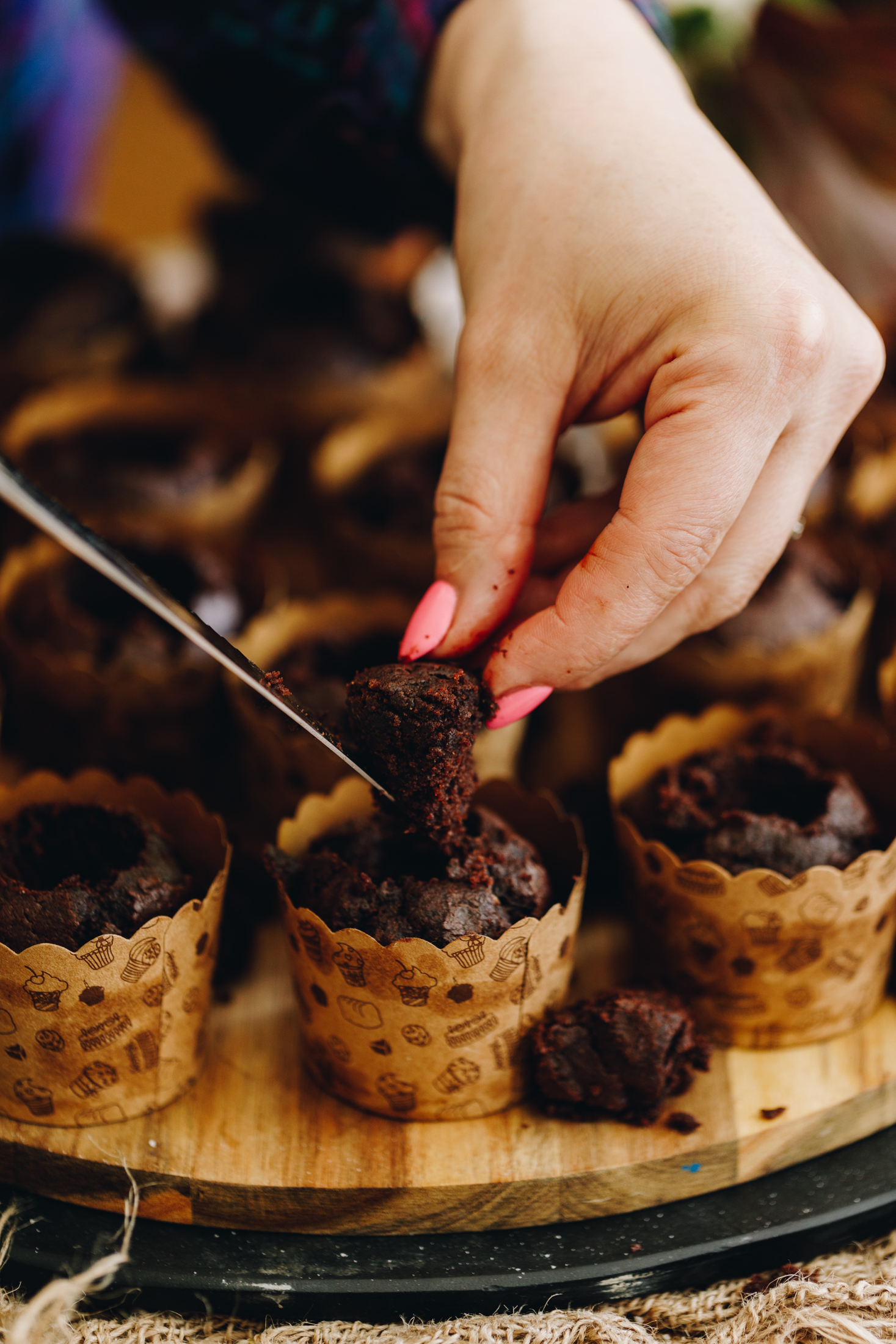 On a round wooden board sits baked chocolate muffins that are in brown cardboard cupcakes cases. They have the centres removed out of them. Naomi is removing the centre out of the centre cupcake.