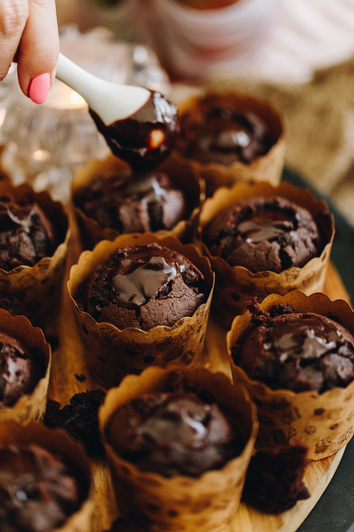 On a round wooden board sits baked chocolate muffins that are in brown cardboard cupcakes cases. They have the centres removed out of them. The holes have been filled with chocolate ganache and Naomi is using a ceramic spoon to add ganache in to one of the muffins.