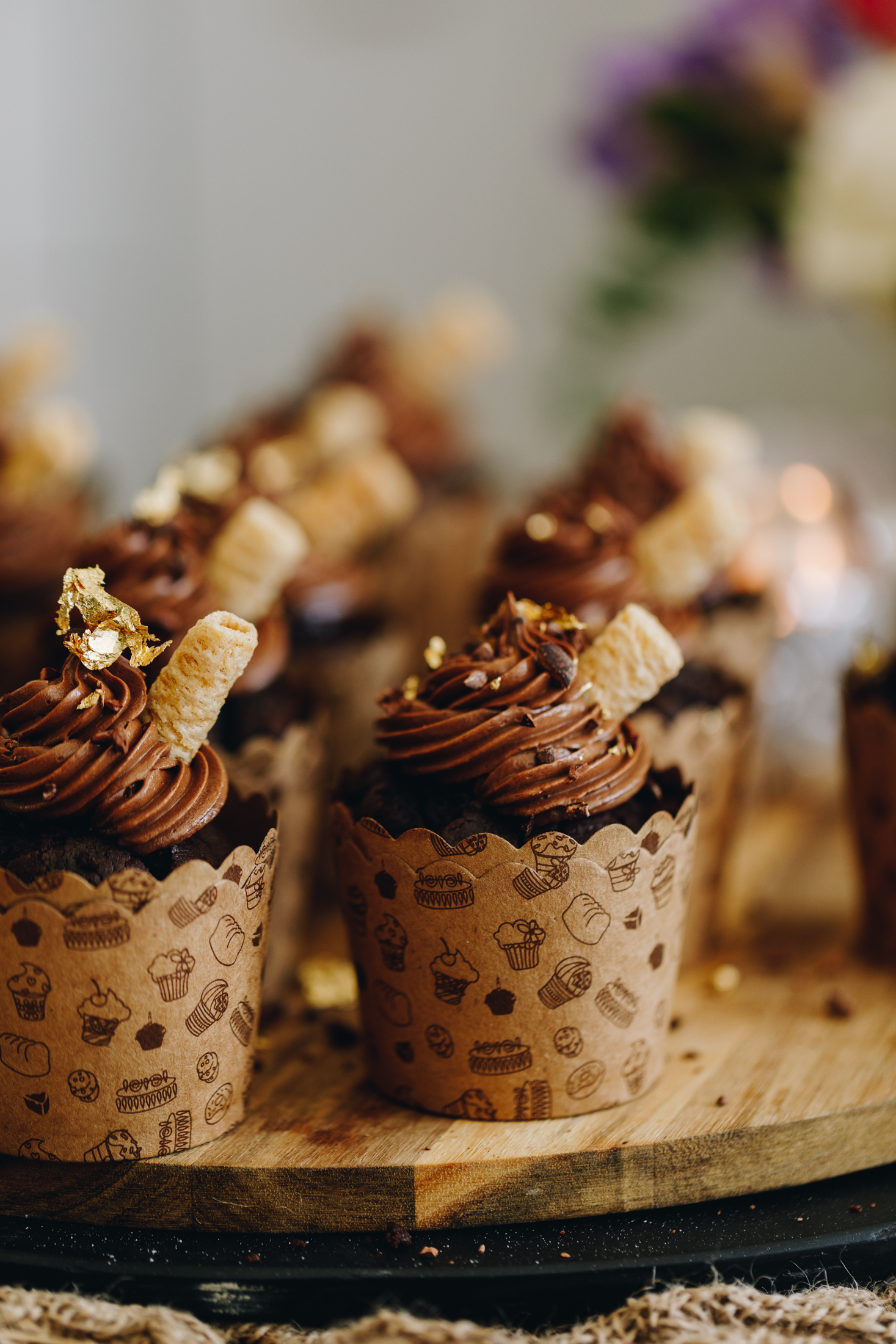 On a round wooden board sits baked chocolate ganache muffins that are in brown cardboard cupcakes cases. They have piped chocolate buttercream on top with a wafer and gold leaf on top of each one. 