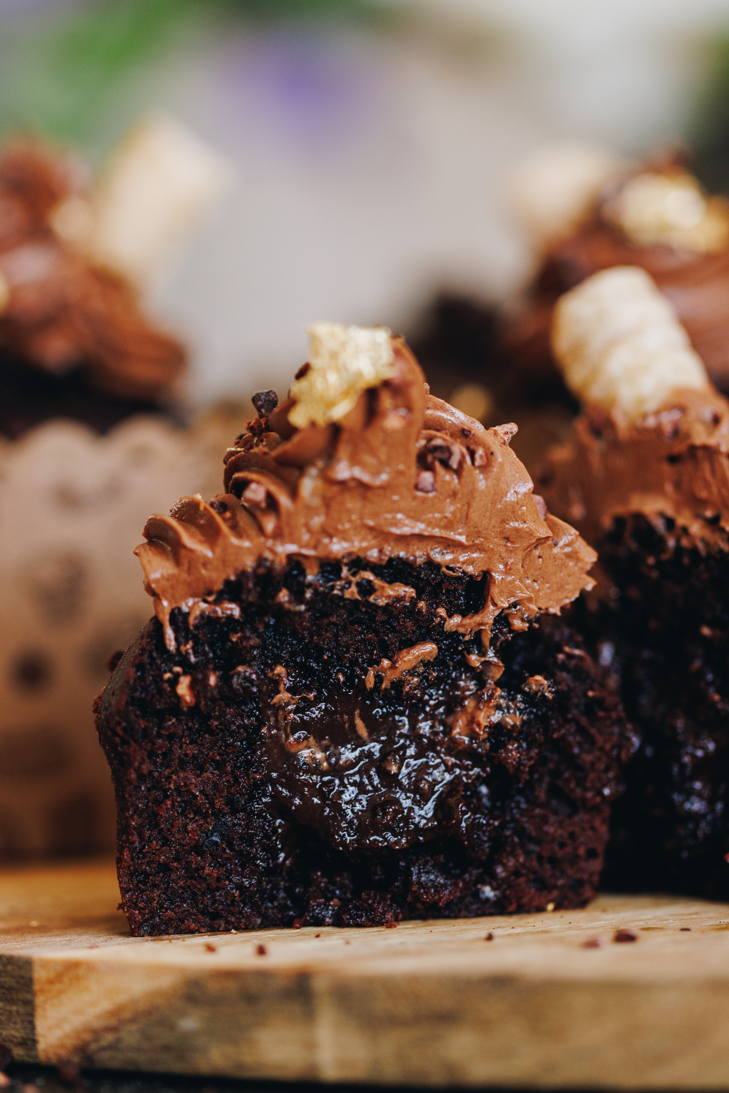 Chocolate ganache muffins sit on a wooden board. One of the muffins is in focus and it has been cut in half to reveal the gooey chocolate centre and the chocolate frosting piled on top.