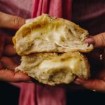 Two cheesy no-knead bread buns are stacked on top of each other and are held by hands. Gooey cheese is oozing out of the fluffy buns.