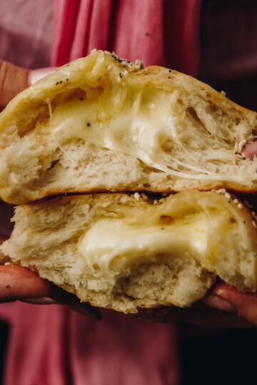 Two cheesy no-knead bread buns are stacked on top of each other and are held by hands. Gooey cheese is oozing out of the fluffy buns.
