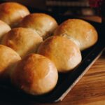 frehsly baked no-knead bread buns are on a black tray, sitting on a wooden table. They are glazed with melted butter.