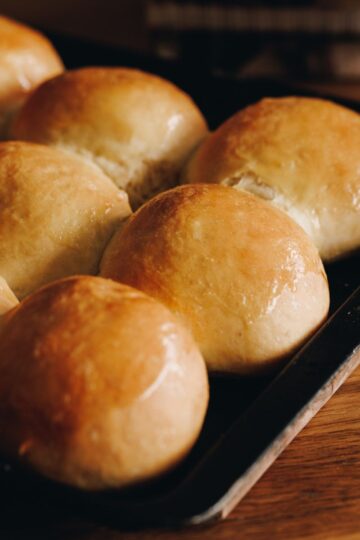 frehsly baked no-knead bread buns are on a black tray, sitting on a wooden table. They are glazed with melted butter.