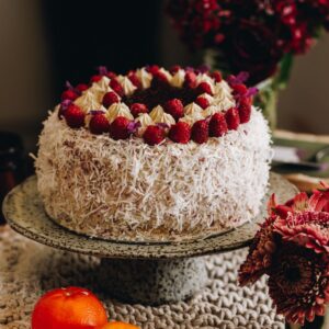 A raspberry lamington cake is on a ceramic cake stand it is covered is shredded coconut and has raspberries around the top with raspberry jam in the centre.