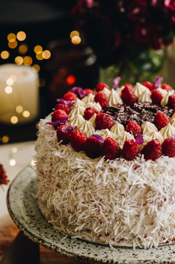 A medium close of a Raspberry Lamington Cake is on a ceramic cake stand. It is decorated in shredded coconut, fresh raspberries around the top, and raspberry jam in the centre.