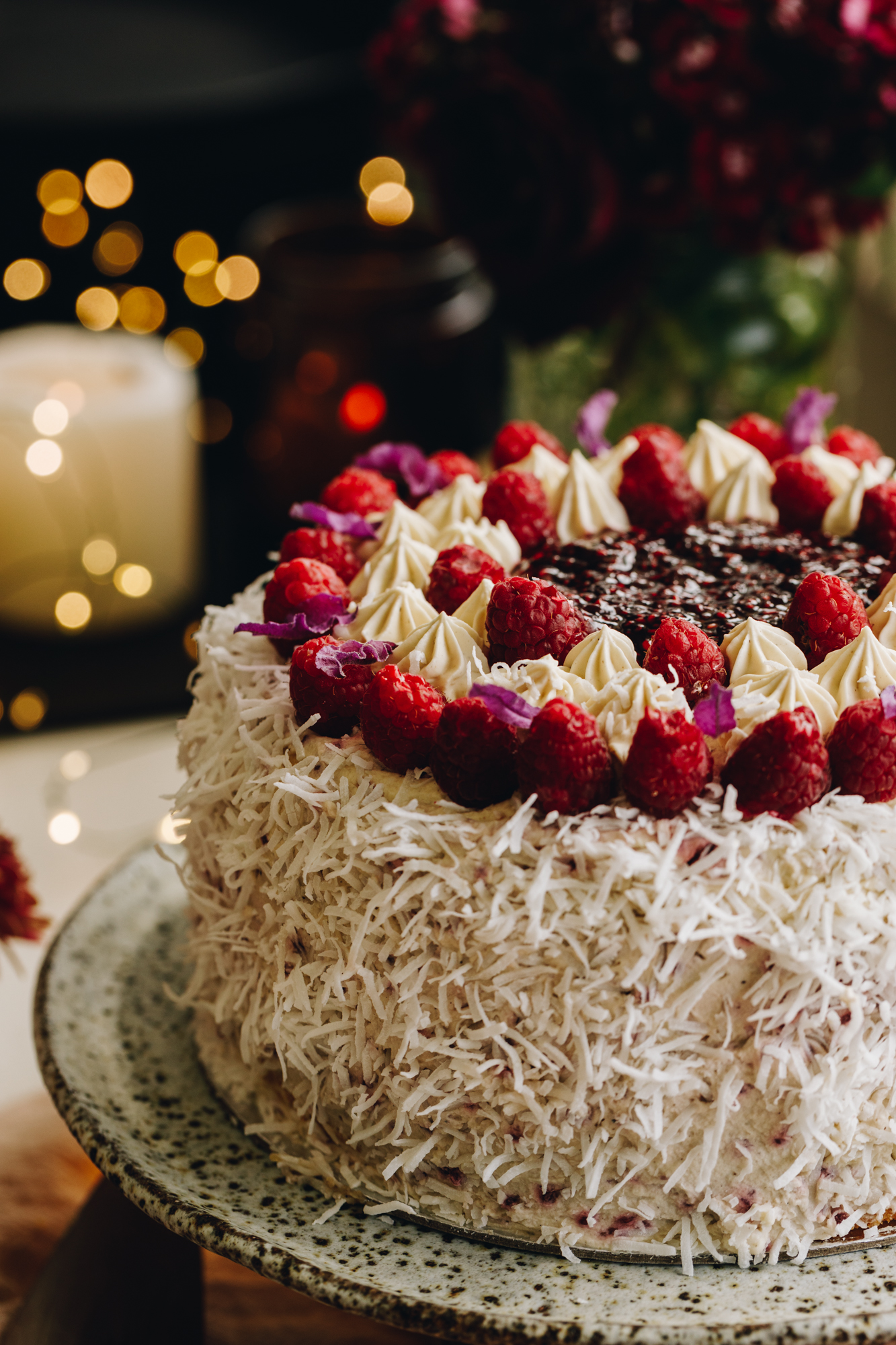 A medium close of a Raspberry Lamington Cake is on a ceramic cake stand. It is decorated in shredded coconut, fresh raspberries around the top, and raspberry jam in the centre.