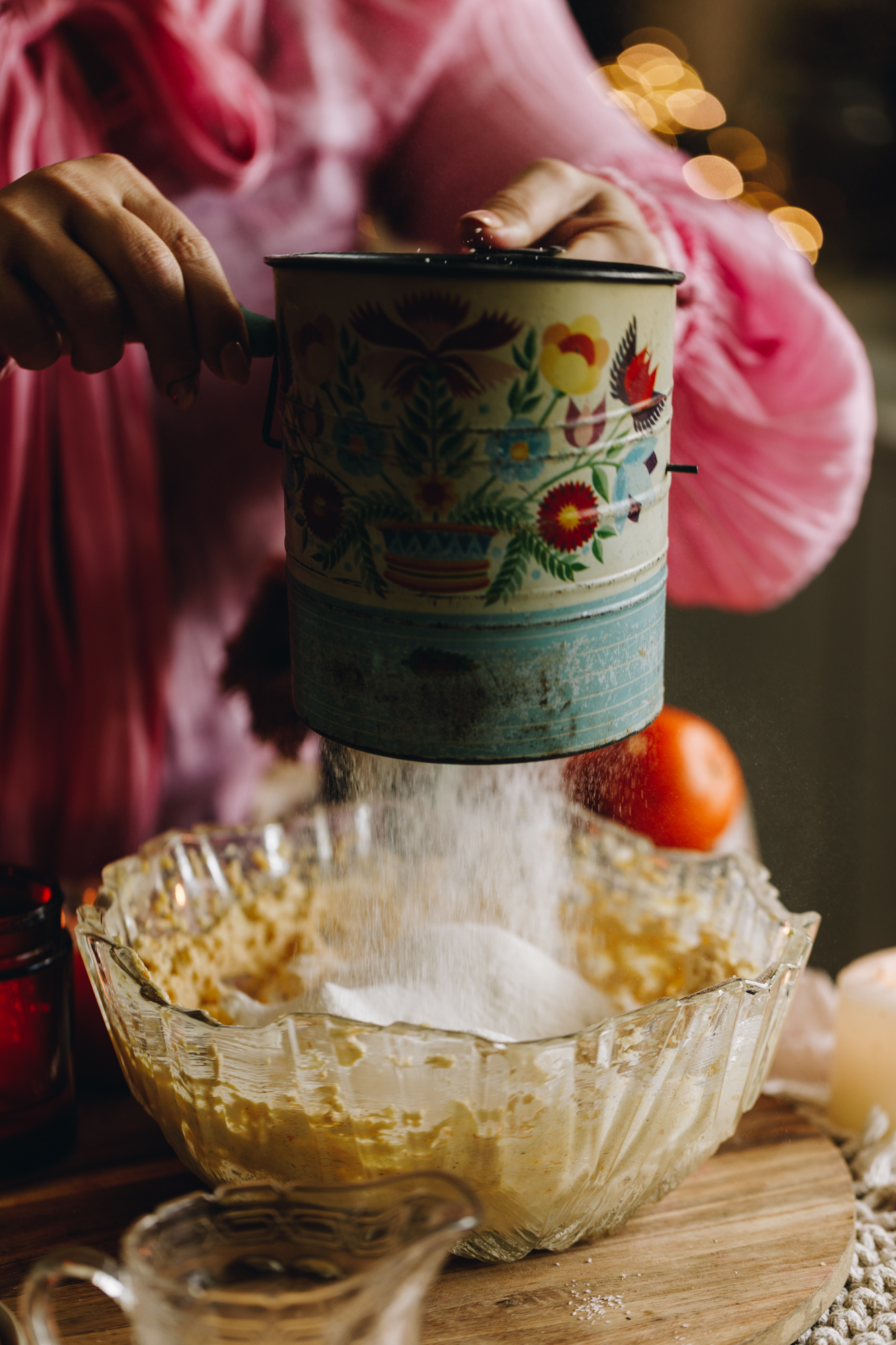An old vintage sifted with a vintage flower pattern is sifting flour in to the whipped sugar and butter mixture, it is on a wooden table with hands turning the wooden handle.