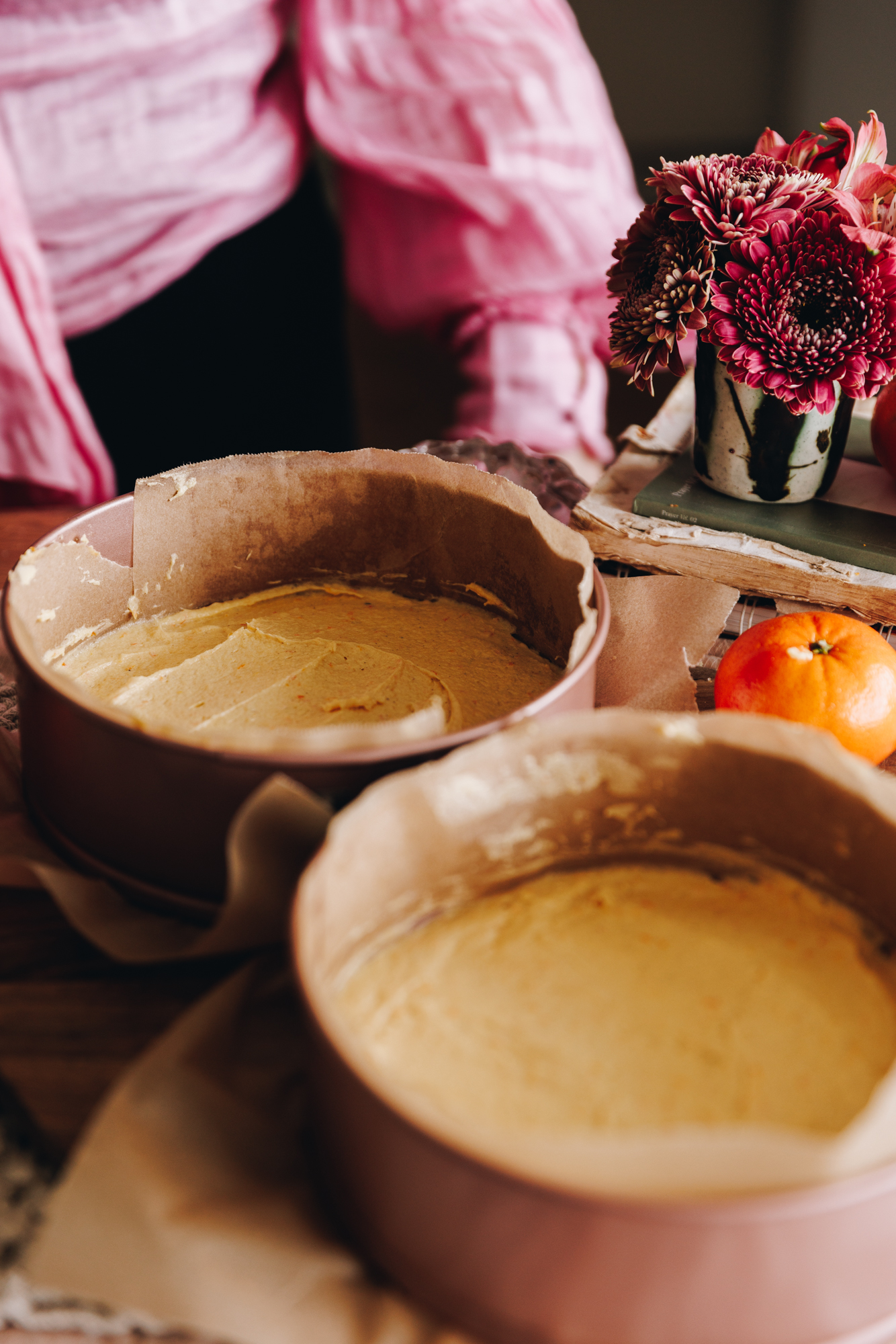 Two pink cake tins have unbaked cake batter in them, they are spread flat and sit on a table. Flowers and a mandarin are in the background.
