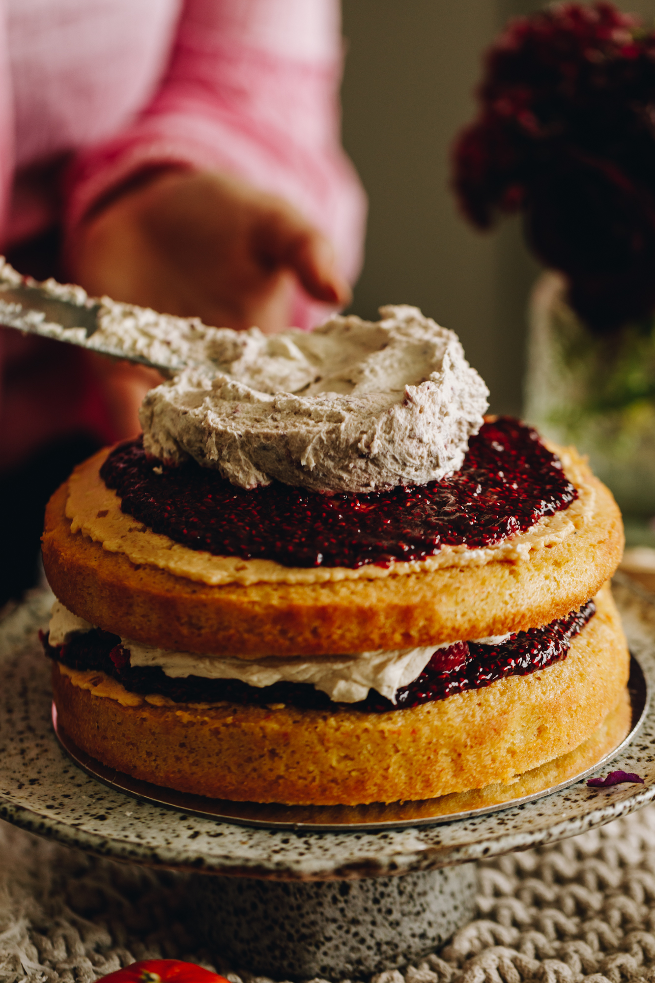 The stable whipped cream is being spread on to the layered sponges of the Raspberry Lamington Cake. The creams being added on top of raspberry jam and the cake is on a ceramic cake stand.