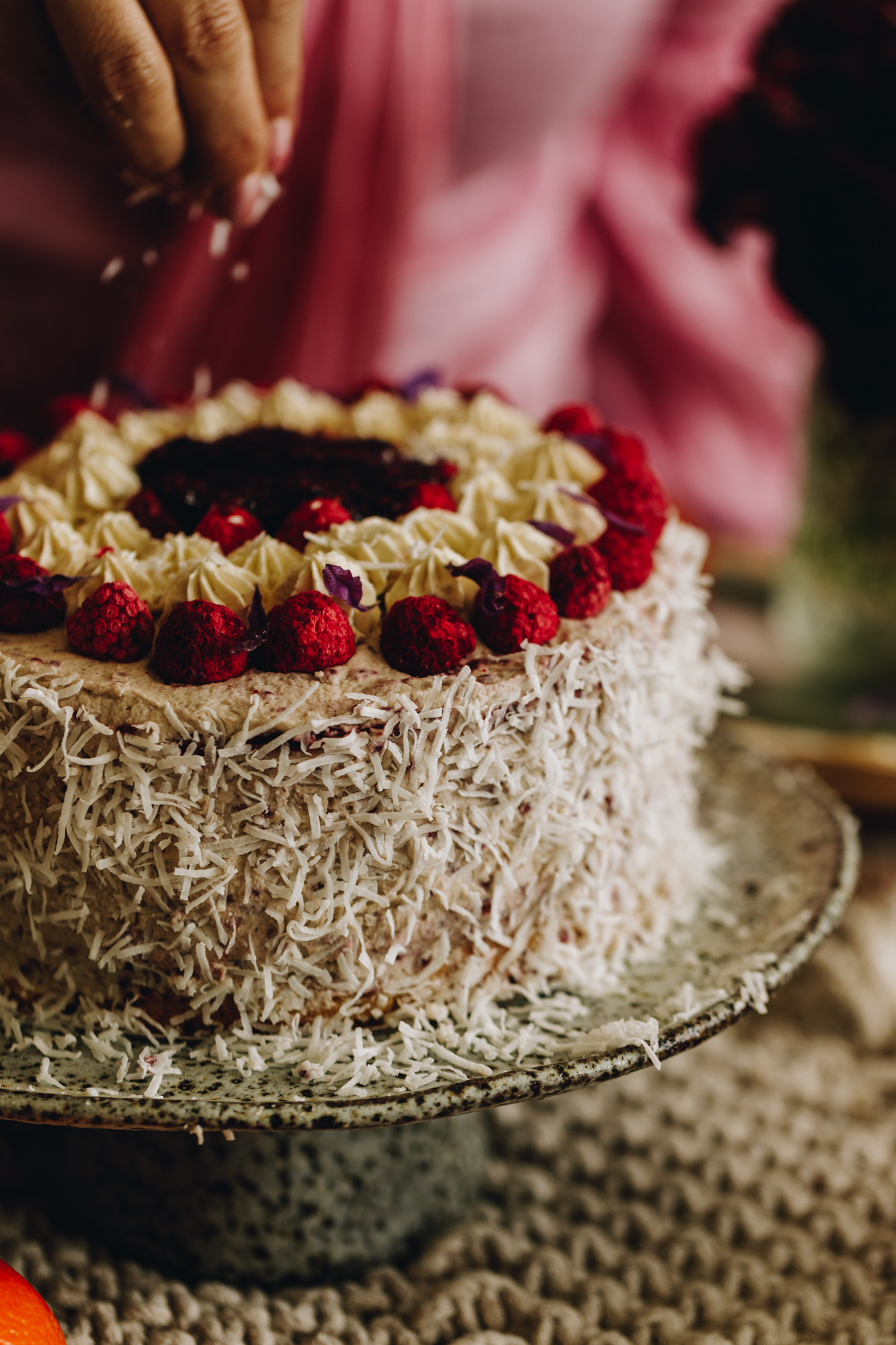 A medium close of a Raspberry Lamington Cake is on a ceramic cake stand. It is being decorated in shredded coconut. Fresh raspberries around the top, and raspberry jam in the centre.