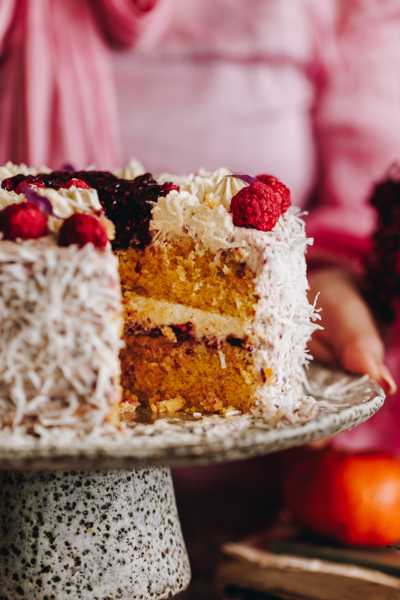 Raspberry Lamington Cake is on a ceramic cake stand. It has been decorated in shredded coconut. Fresh raspberries around the top, and raspberry jam in the centre. A slice has been removed revealing the inside layer and texture of the cake.