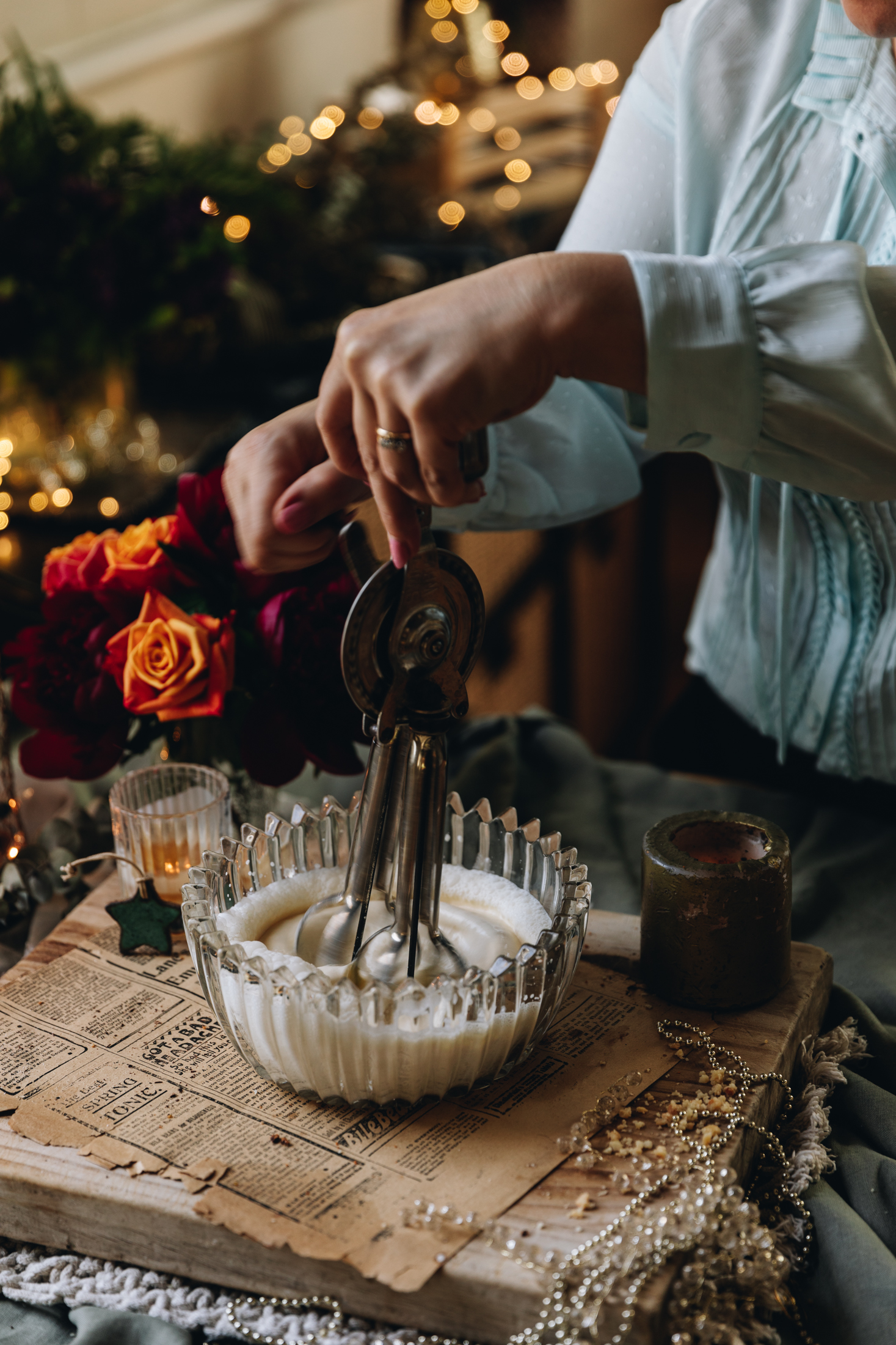 Cream is being whipped in a glass bowl. It is on old newspaper clippings and a wooden board with flowers in the background.