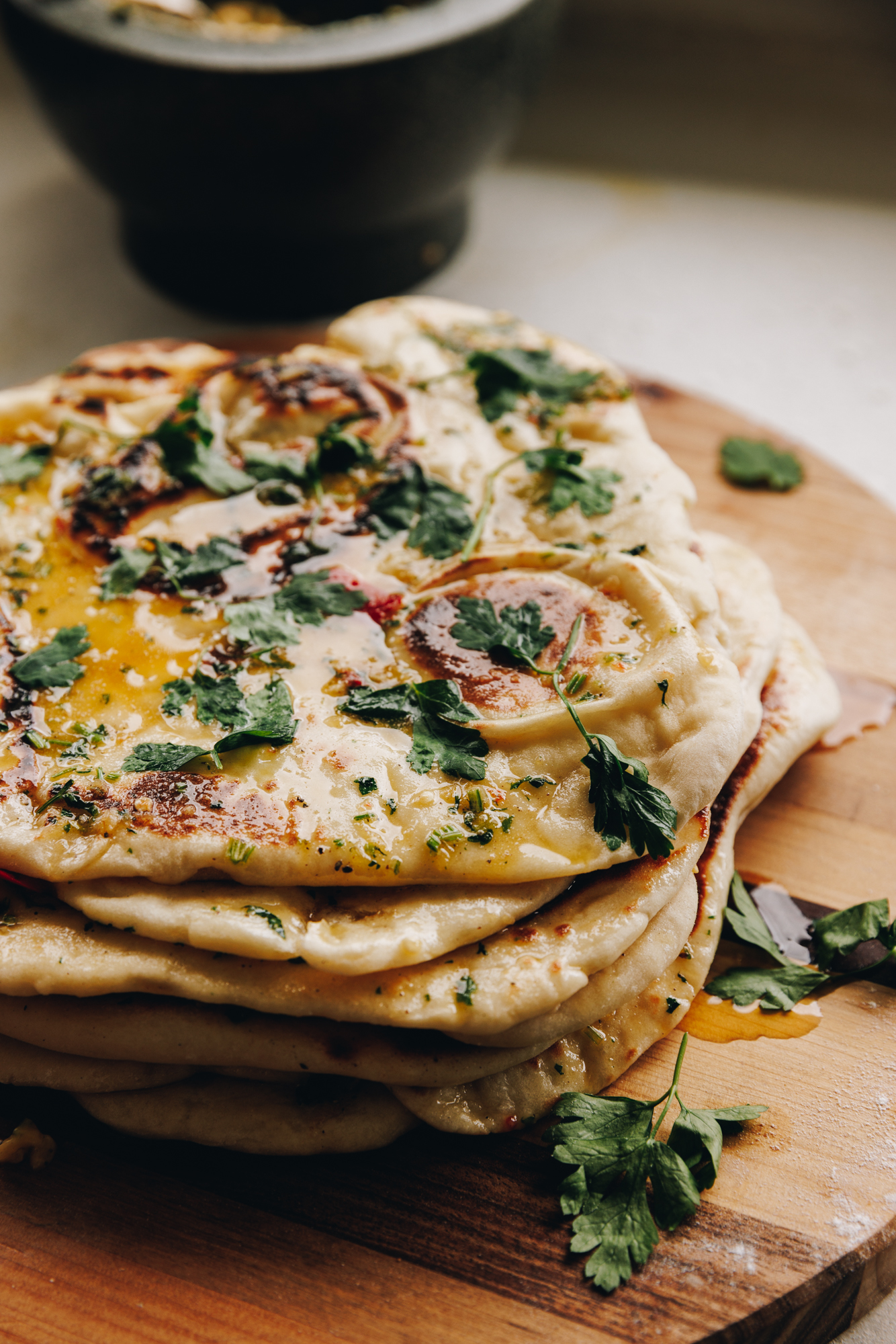 A pile of cooked Garlic Butter Naan Bread sits on a wooden chopping board. The melted garlic butter is visible as are the pieces of fresh Italian parsley. 