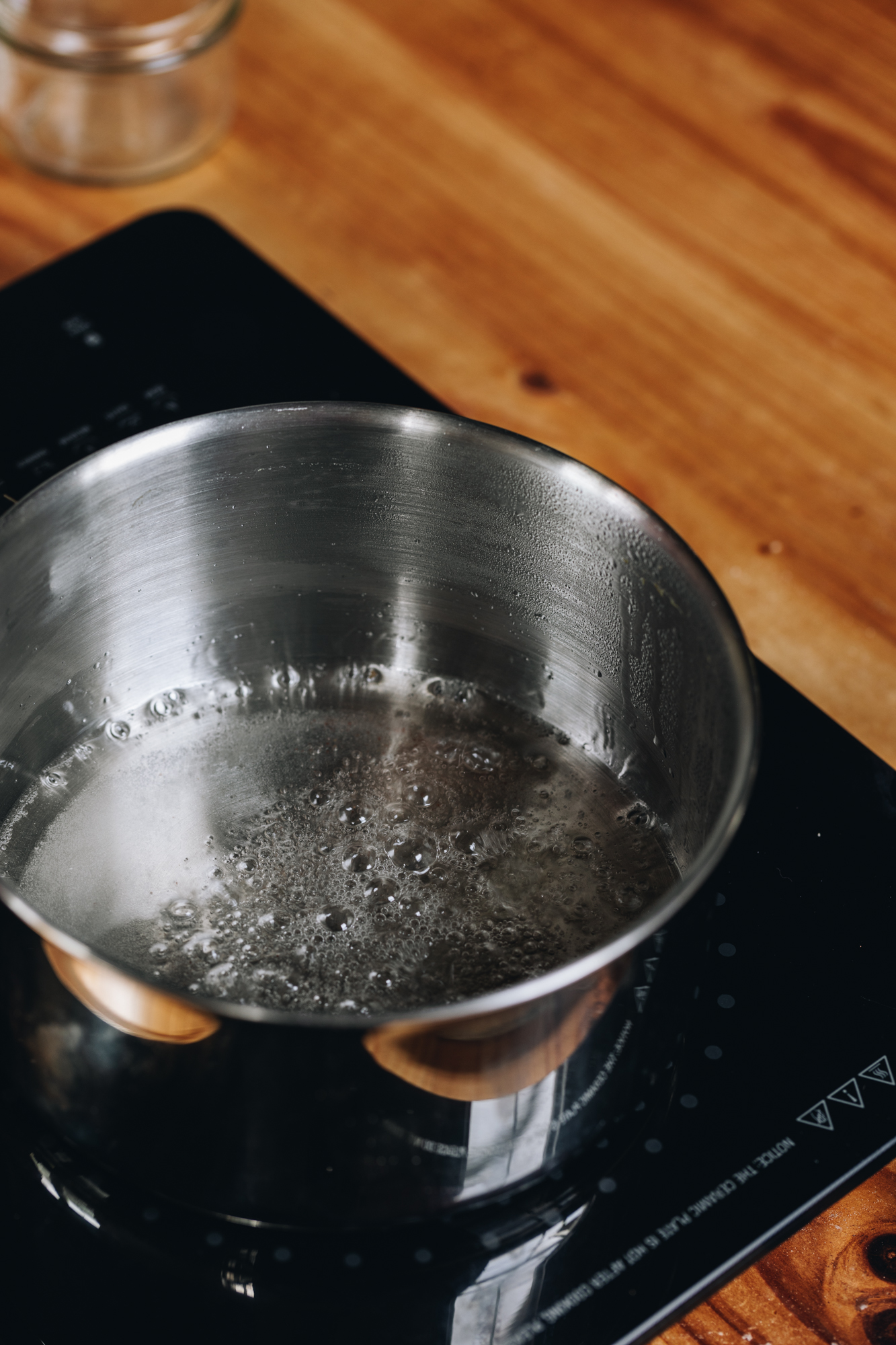 A sugar syrup is boiling in a silver pot on top of a black portable stove. It is on a wooden table. 