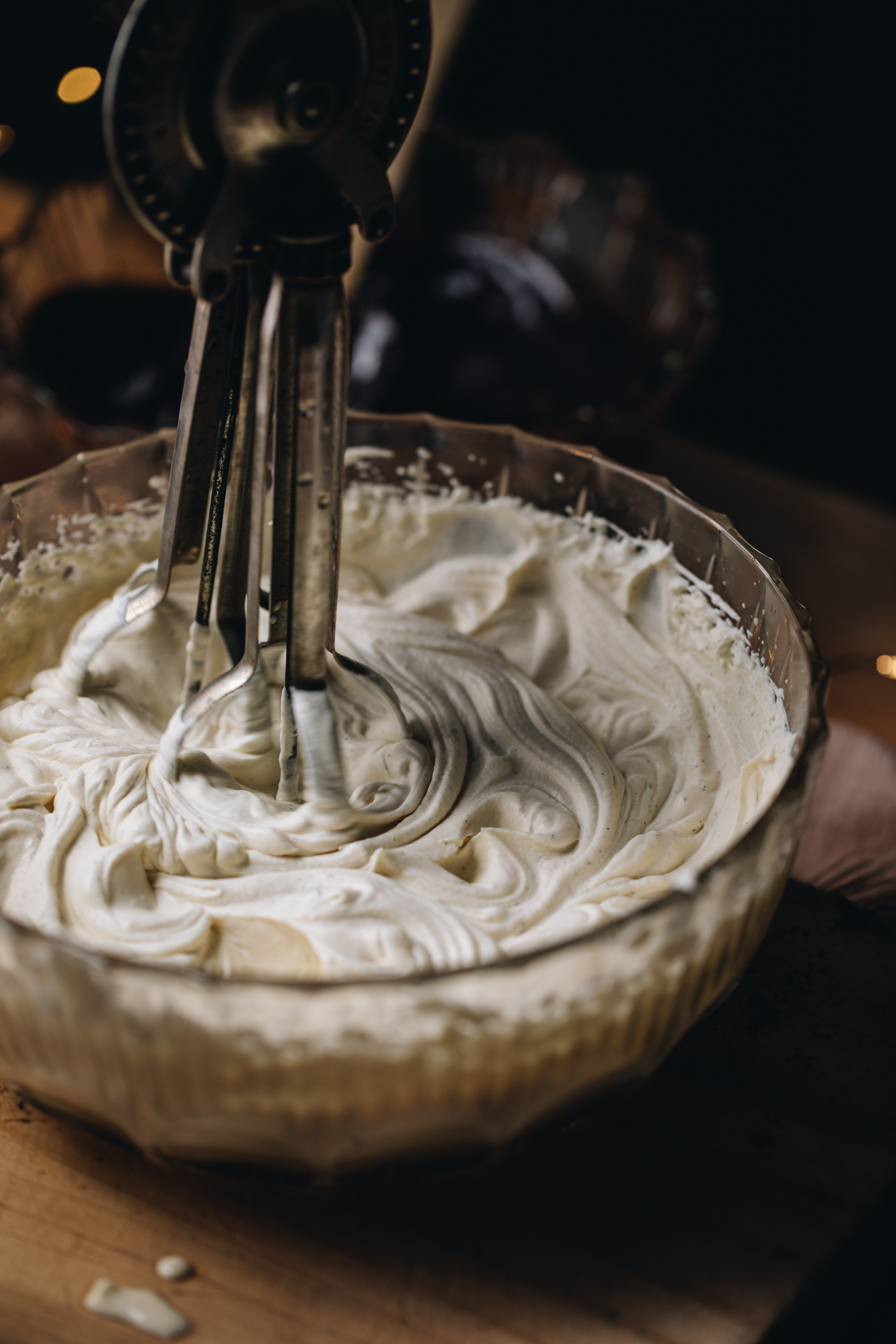 Cream is being whipped in a vintage glass bowl. The cream is thick and ribbony. It is being whipped by a vintage egg beater on a wooden table.